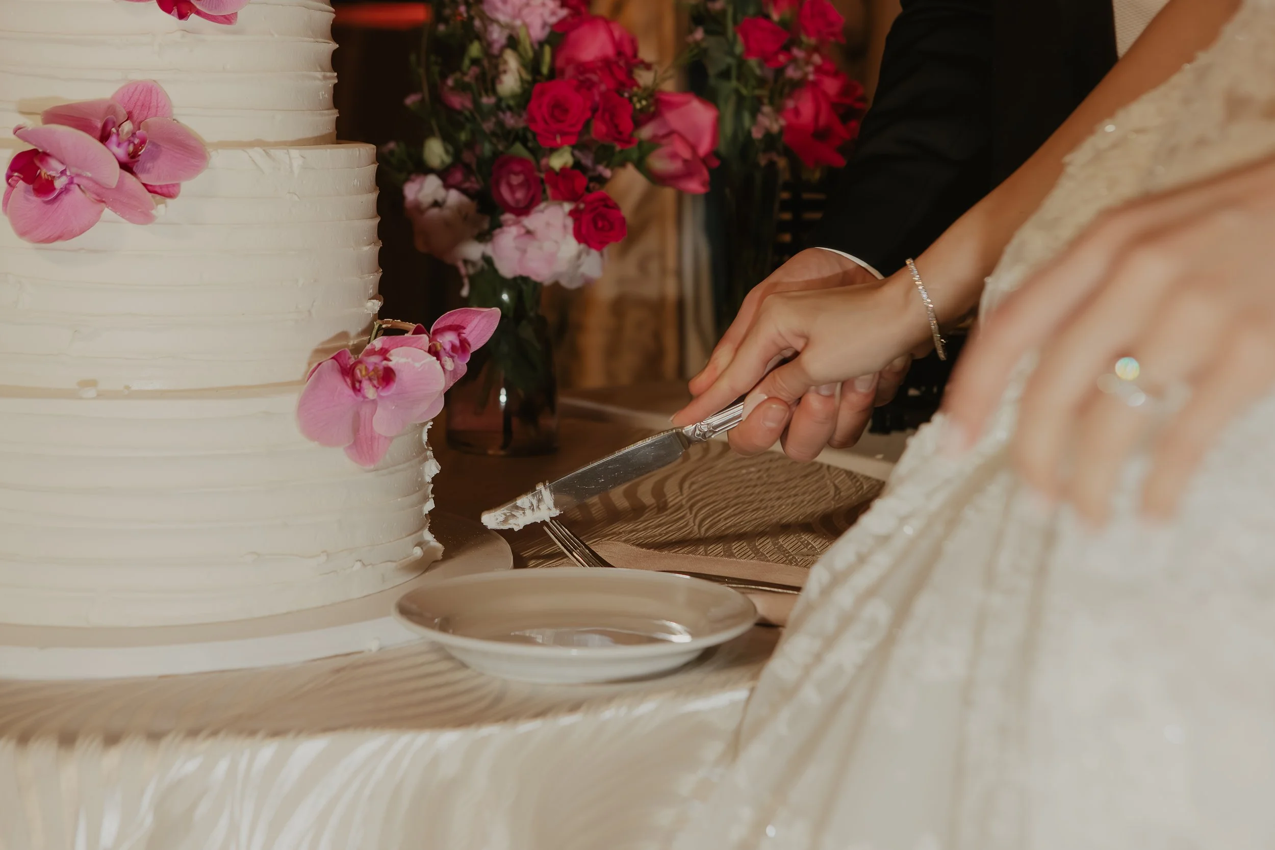A wedding cake decorated with pink orchids on a table, with a bride and groom cutting the cake, surrounded by floral arrangements of pink and red roses.