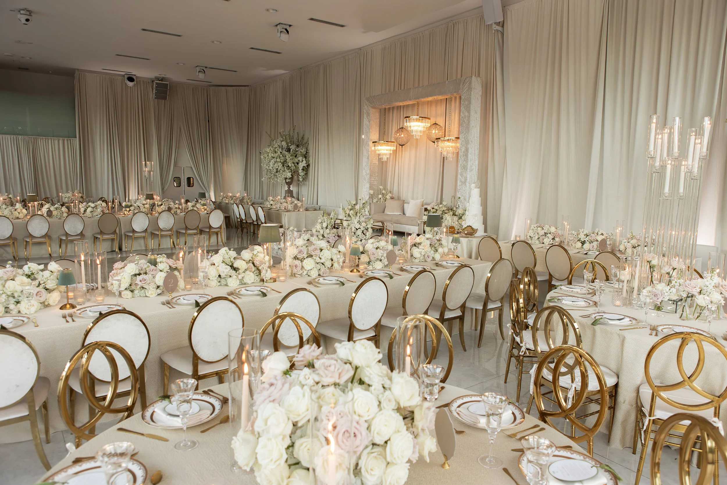Elegant wedding reception decorated with white and blush flowers, tall floral centerpieces, candles, and gold-accented tableware in a bright room with cream drapes and chandelier lighting.