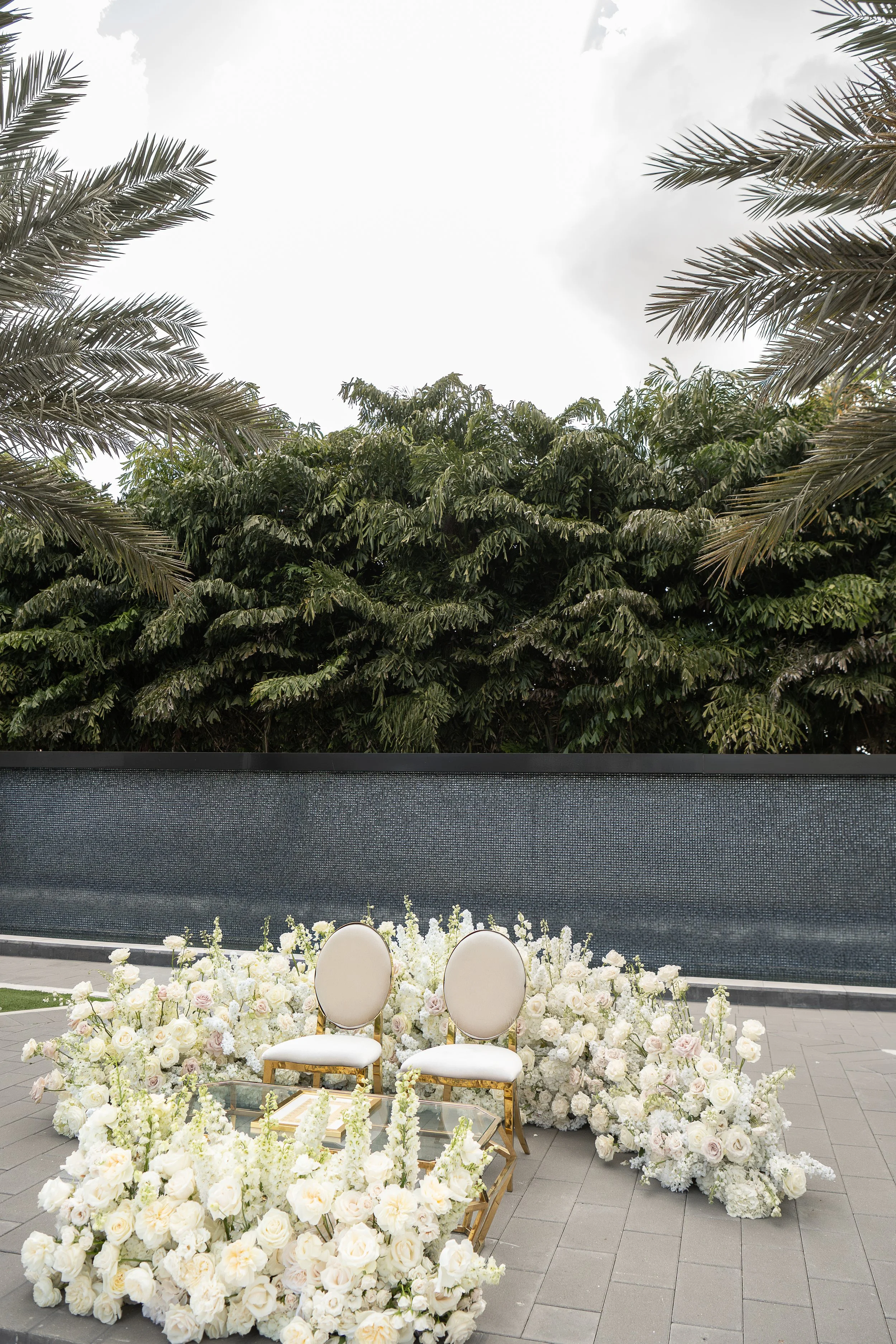 A wedding ceremony setup featuring two white chairs with gold trim in front of an extensive arrangement of white and pale pink flowers, with lush greenery and tall palm trees in the background.