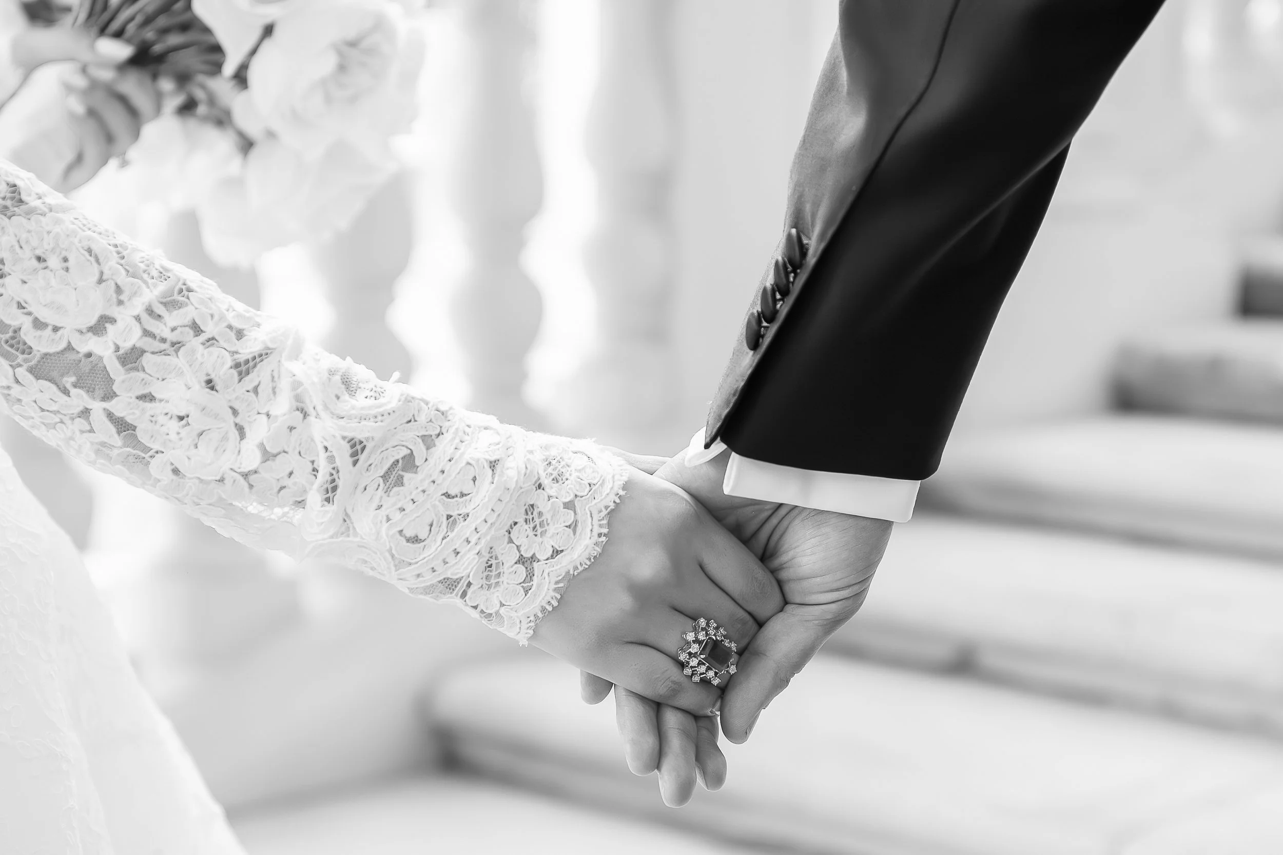 Close-up of a couple holding hands during a wedding ceremony. The bride's hand is adorned with lace sleeves and rings, while the groom's hand is in a suit with buttons visible on the cuff.