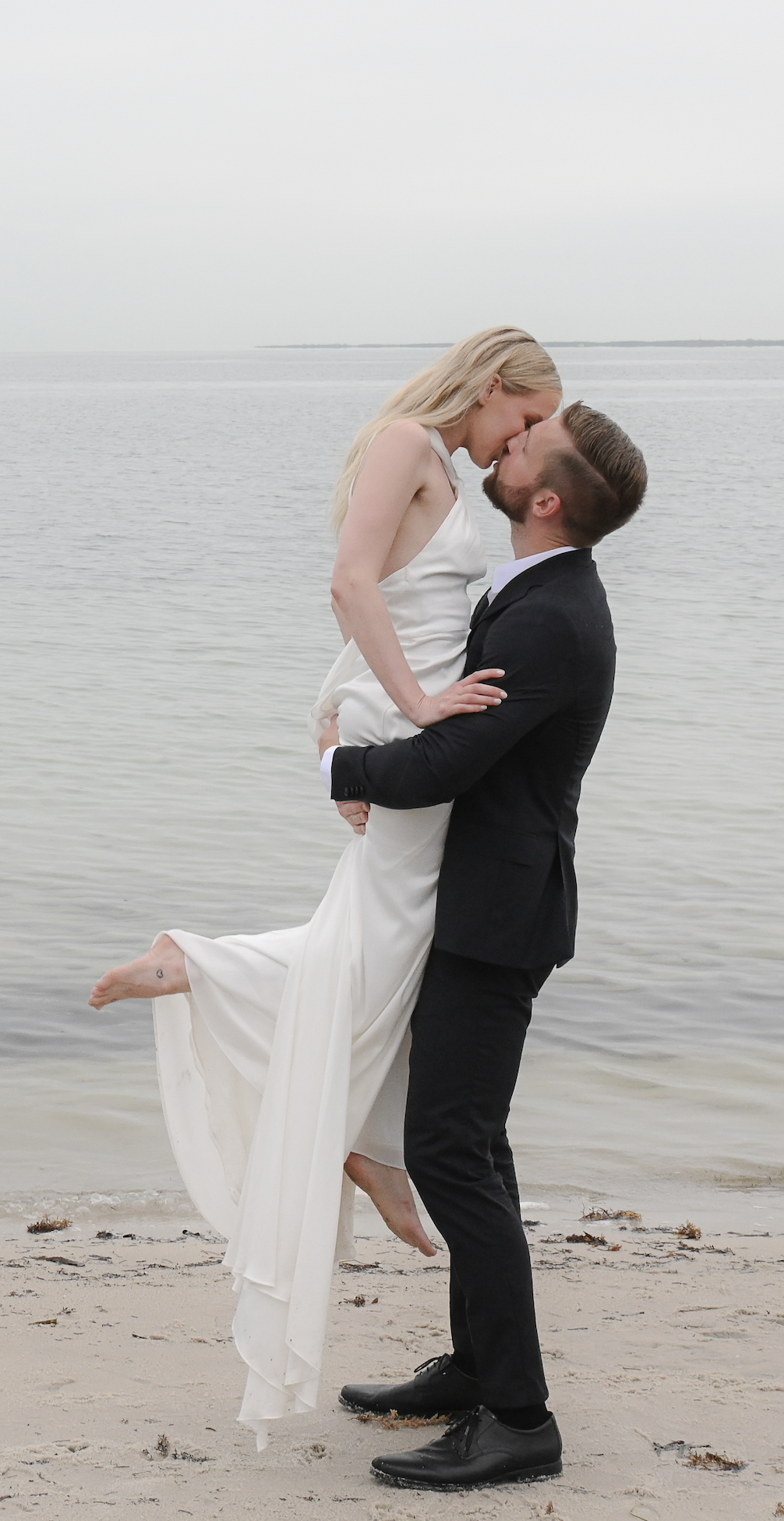 A couple in wedding attire on a beach, with the groom lifting the bride, their faces close together, water in the background.