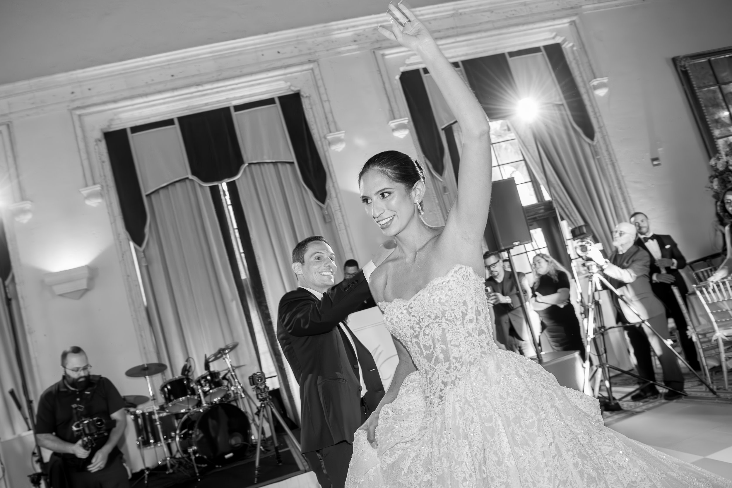 A bride and groom dancing at their wedding reception in an elegant room with tall windows and curtains, surrounded by guests and a live band.