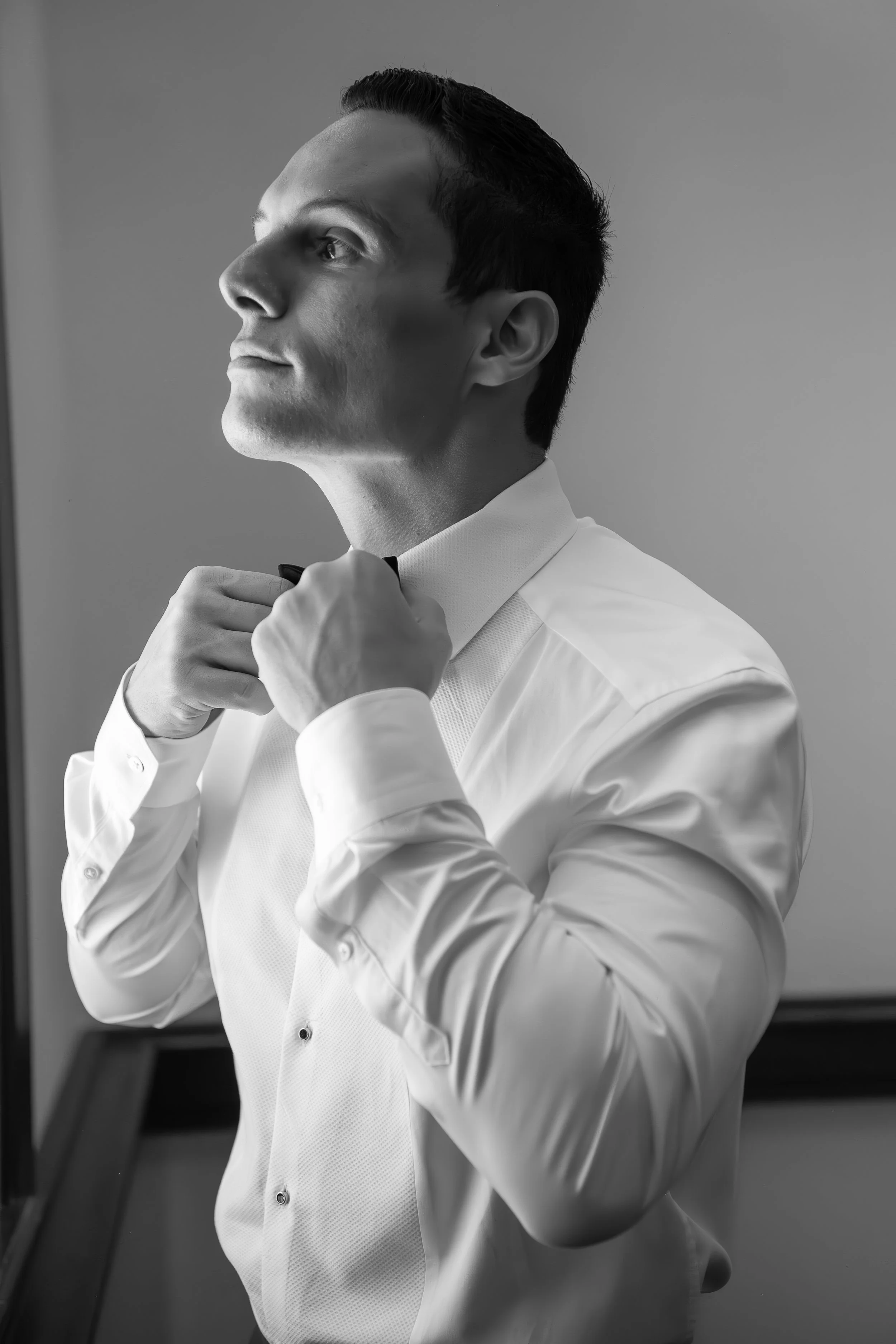 A black and white photo of a man in a formal white shirt adjusting his bow tie, looking slightly upwards.