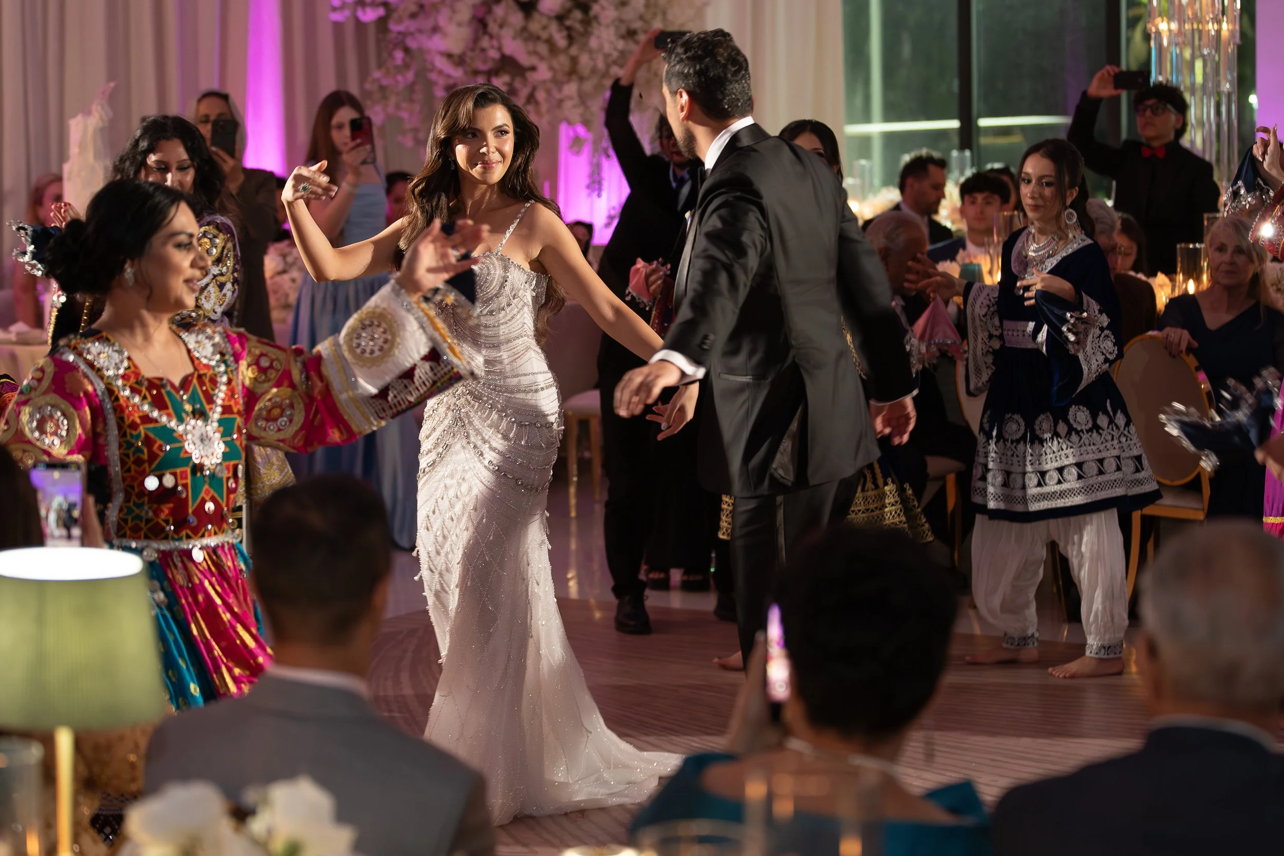 People dancing at a wedding reception indoors, with a woman in a white wedding dress and a man in a black tuxedo dancing in the center, surrounded by guests in colorful traditional attire and formal wear, decorated with pink lighting and floral arran