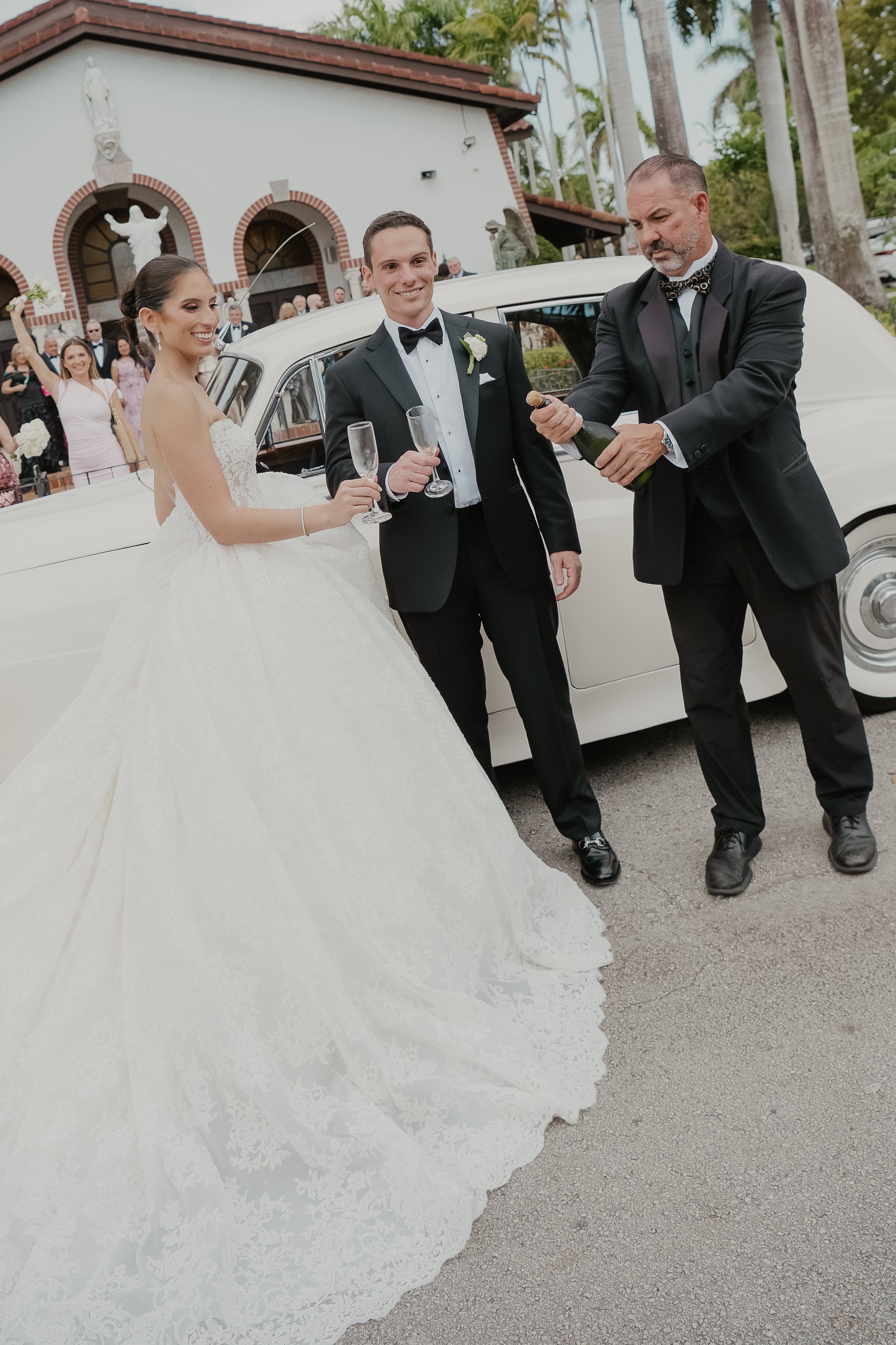 A wedding scene outside a church with a bride and groom, and a man opening champagne bottles while guests watch, some cheering and taking photos.