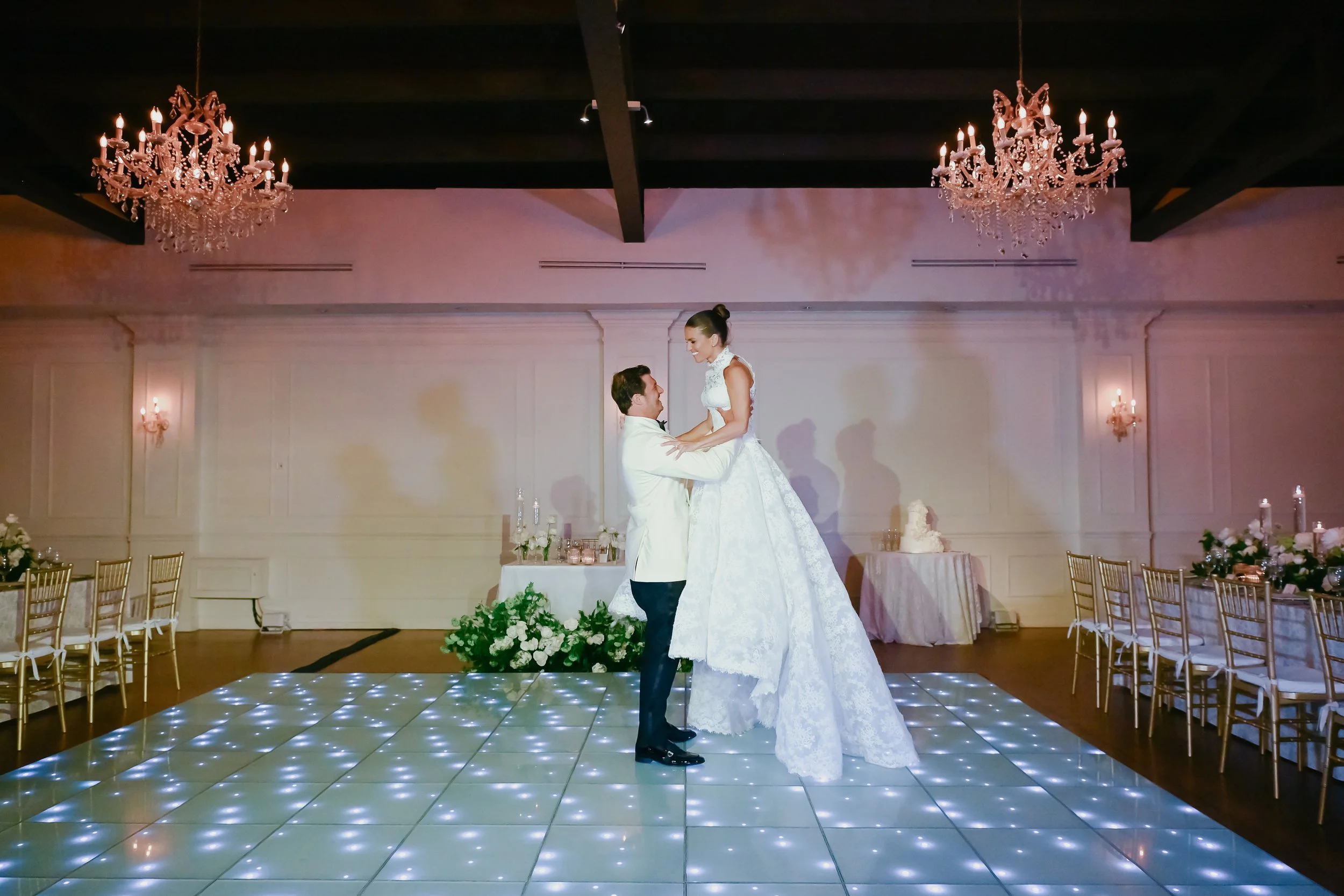 A bride and groom dancing at their wedding reception in a ballroom with chandeliers and decorated tables.