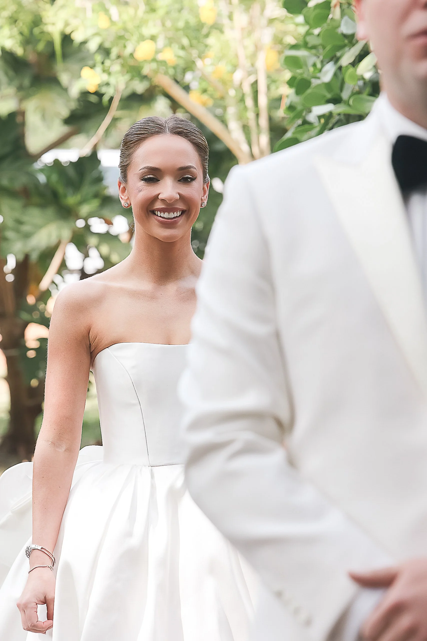 Bride wearing a strapless white wedding dress smiling outdoors, with a groom in a white tuxedo and black bow tie partially visible in the foreground, surrounded by greenery and yellow flowers.