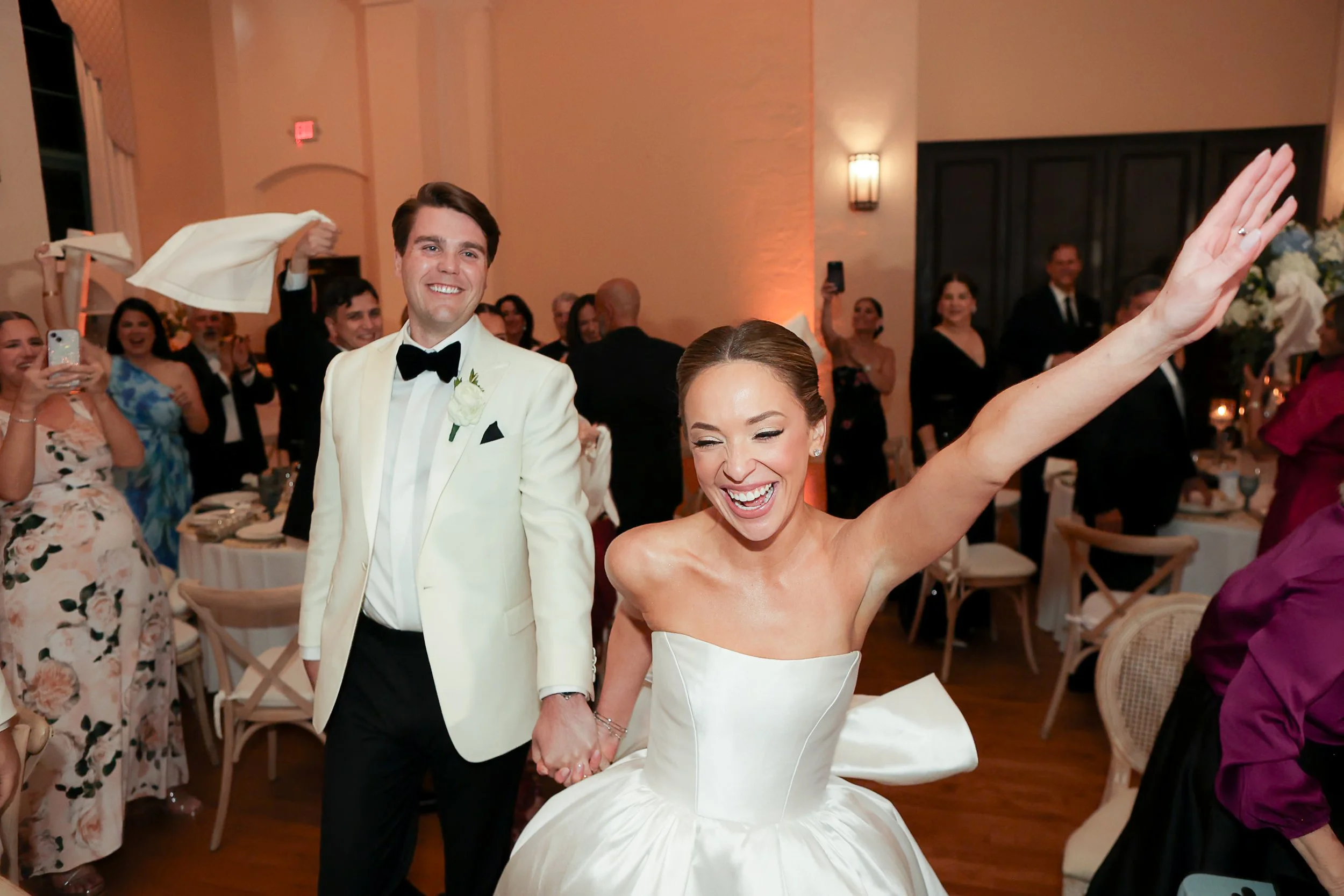 A wedding reception scene with a bride and groom. The groom, in a white tuxedo jacket with a black bow tie, is smiling and holding the bride's hand. The bride is dressed in a white wedding gown, smiling, and raising her hand. Guests are in the backgr