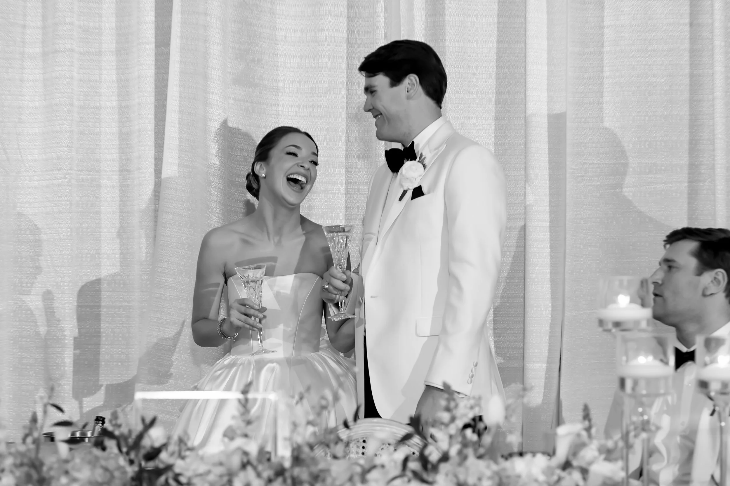 Black and white photo of a wedding reception. A bride in a strapless wedding dress and a groom in a white tuxedo with a black bowtie are smiling and holding champagne glasses. Two guests are partially visible, one sitting at the table and another sta