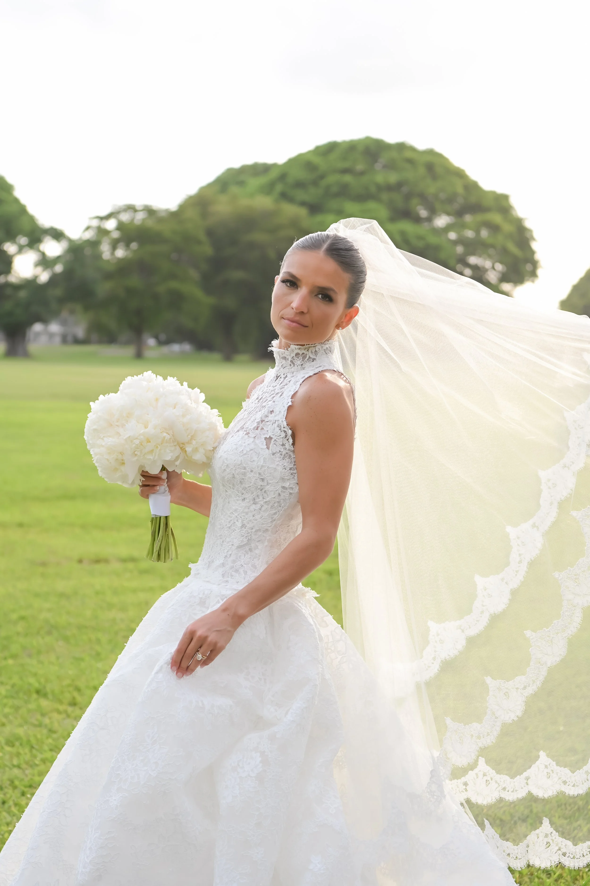 A bride in a white lace wedding dress holding a bouquet of white flowers outdoors with trees and grass in the background.