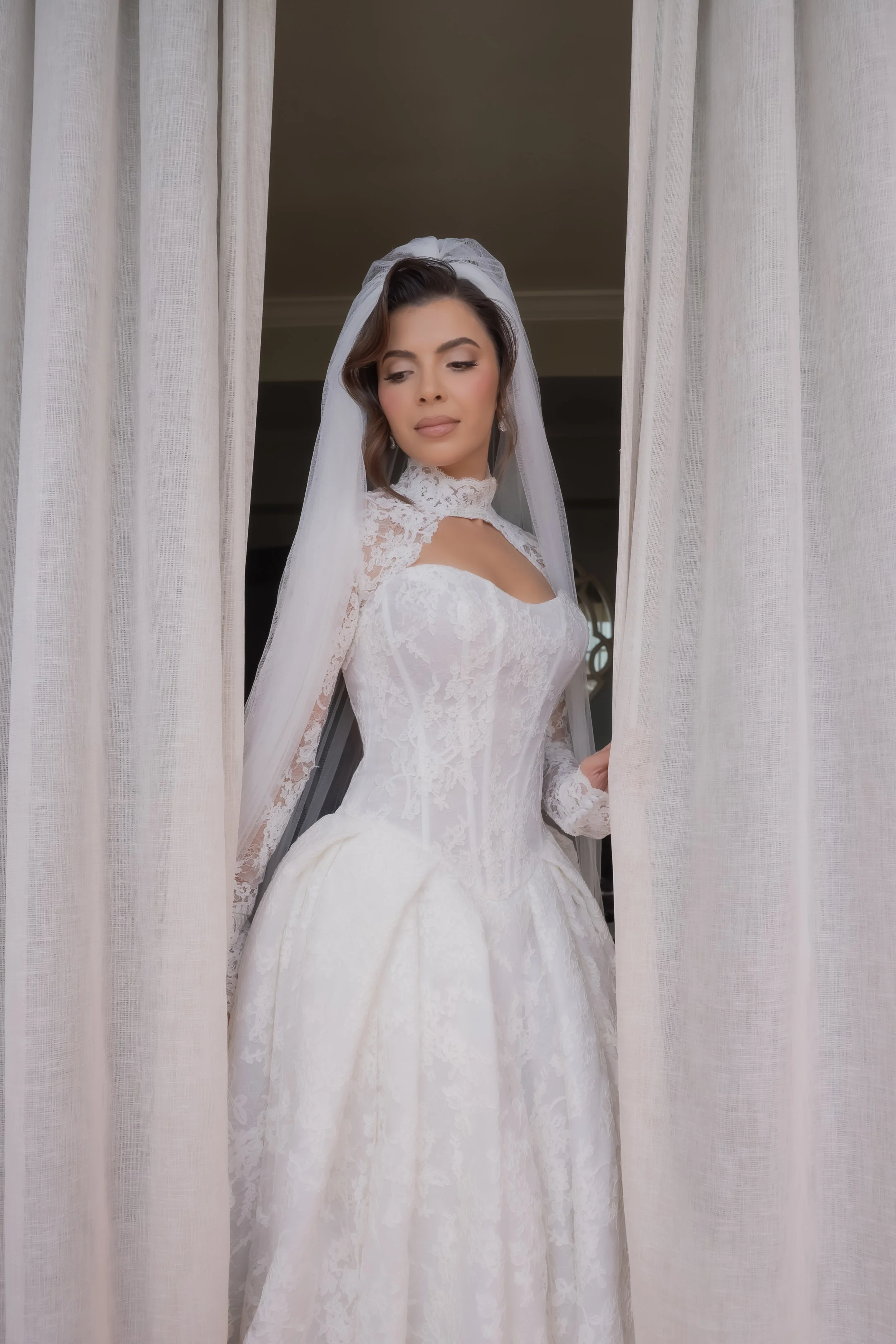 A bride in a white lace wedding dress and veil standing between beige curtains, looking thoughtfully downward.