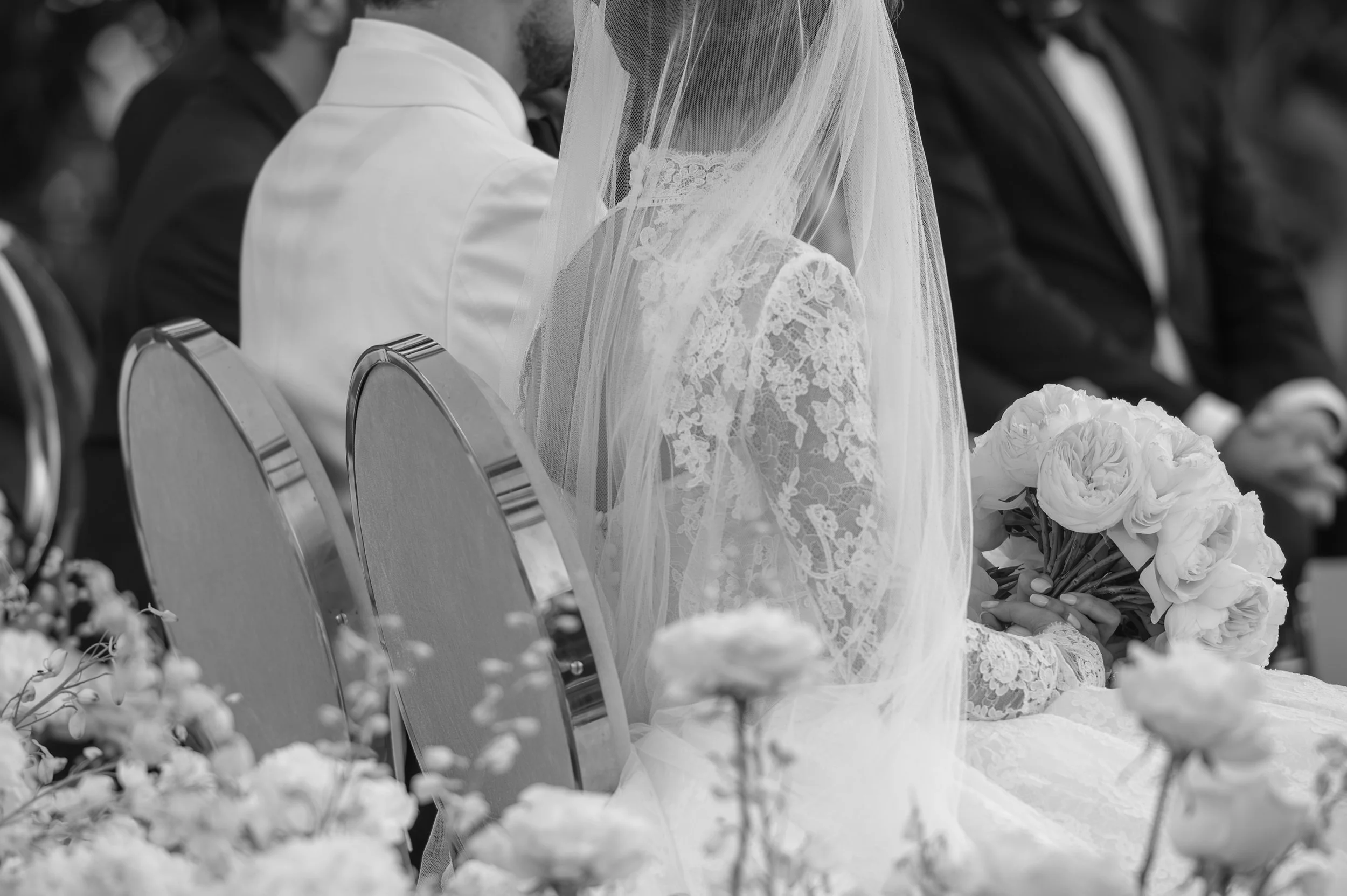 Black-and-white photo of a bride holding a bouquet of white flowers, sitting among chairs at a wedding ceremony. She is wearing a lace wedding dress and a veil. Other guests are seated nearby.