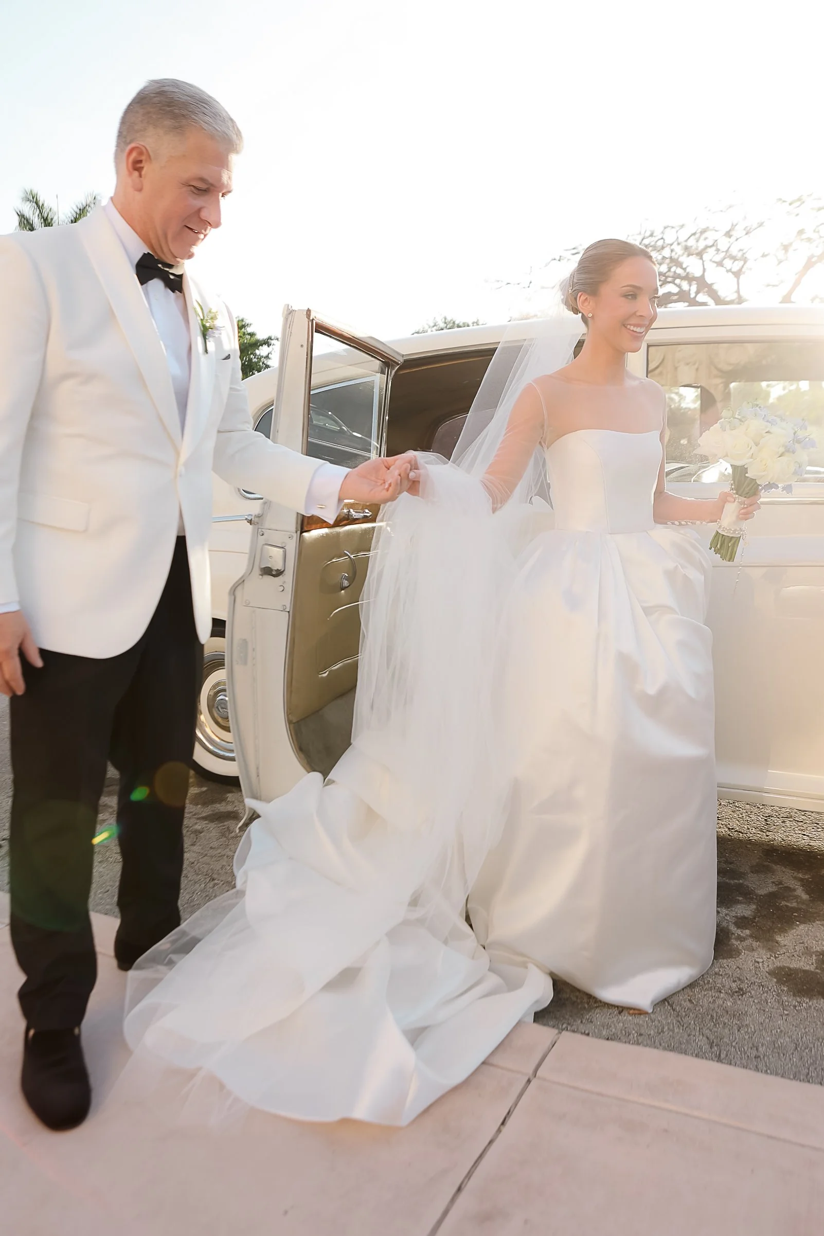 A bride in a white wedding gown holding a bouquet of flowers, exiting a vintage limousine car, with a man in a tuxedo holding her hand.
