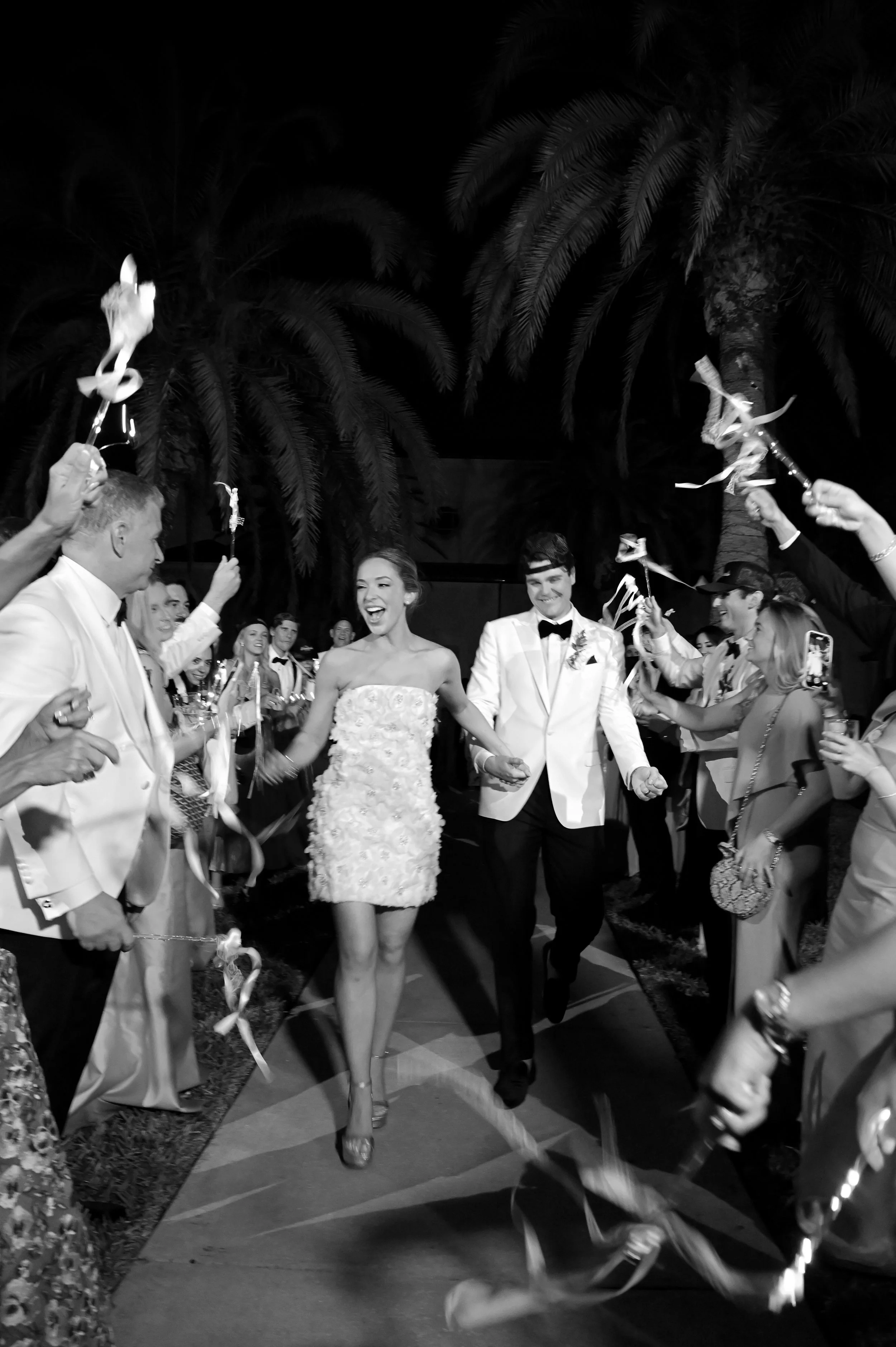 A wedding reception with guests celebrating, holding ribbon wands, with a bride and groom walking down a decorated aisle outdoors at night.