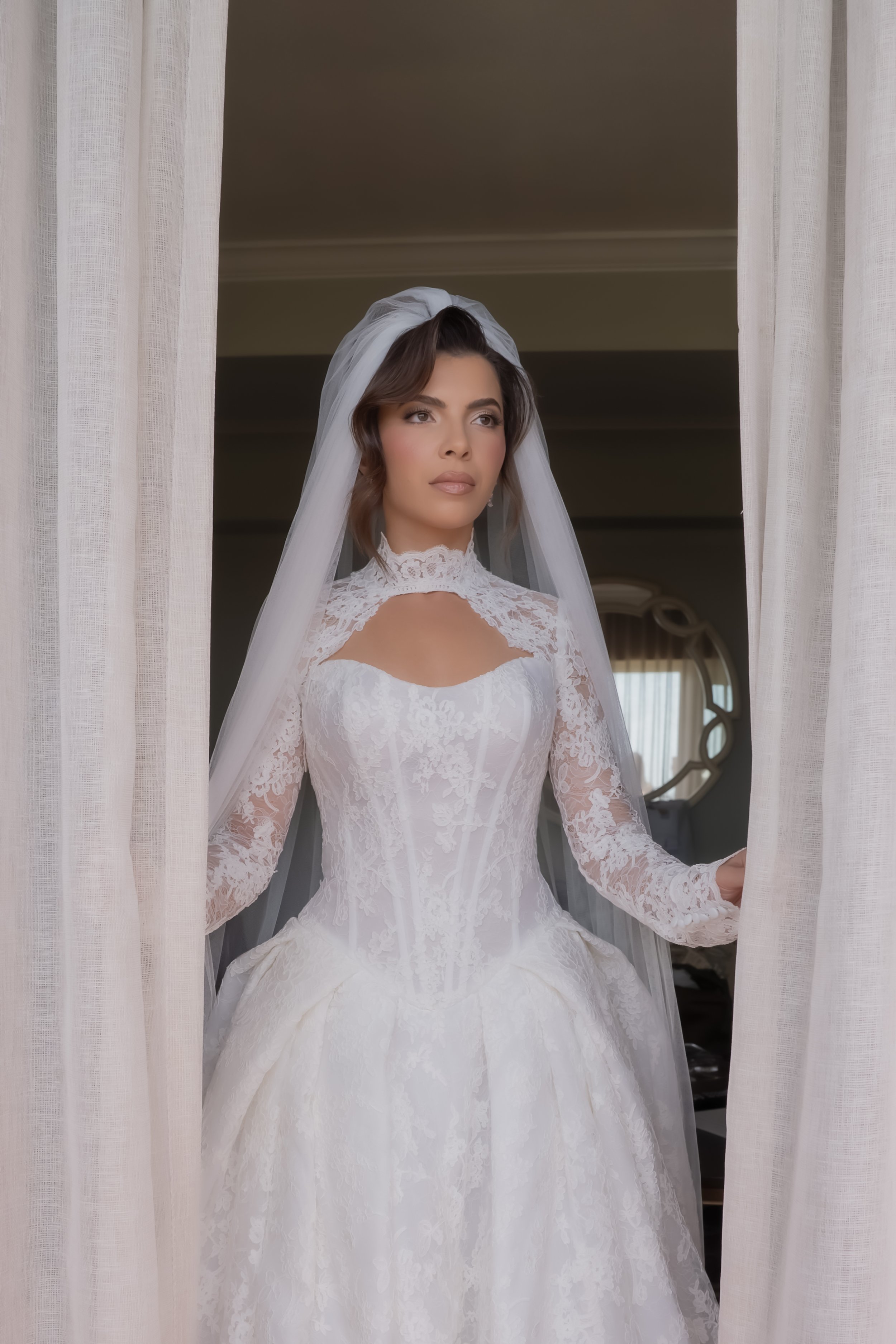 A woman in a white bridal gown standing behind curtains, looking out the window.
