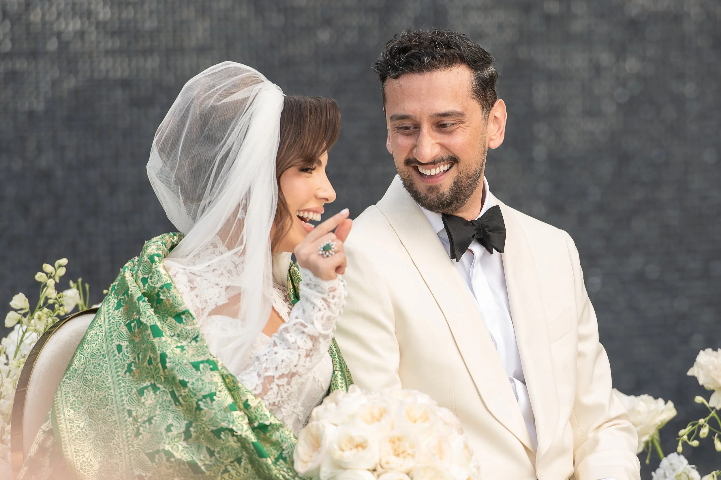 A bride and groom smiling at each other at their wedding reception, with flowers and a dark background.