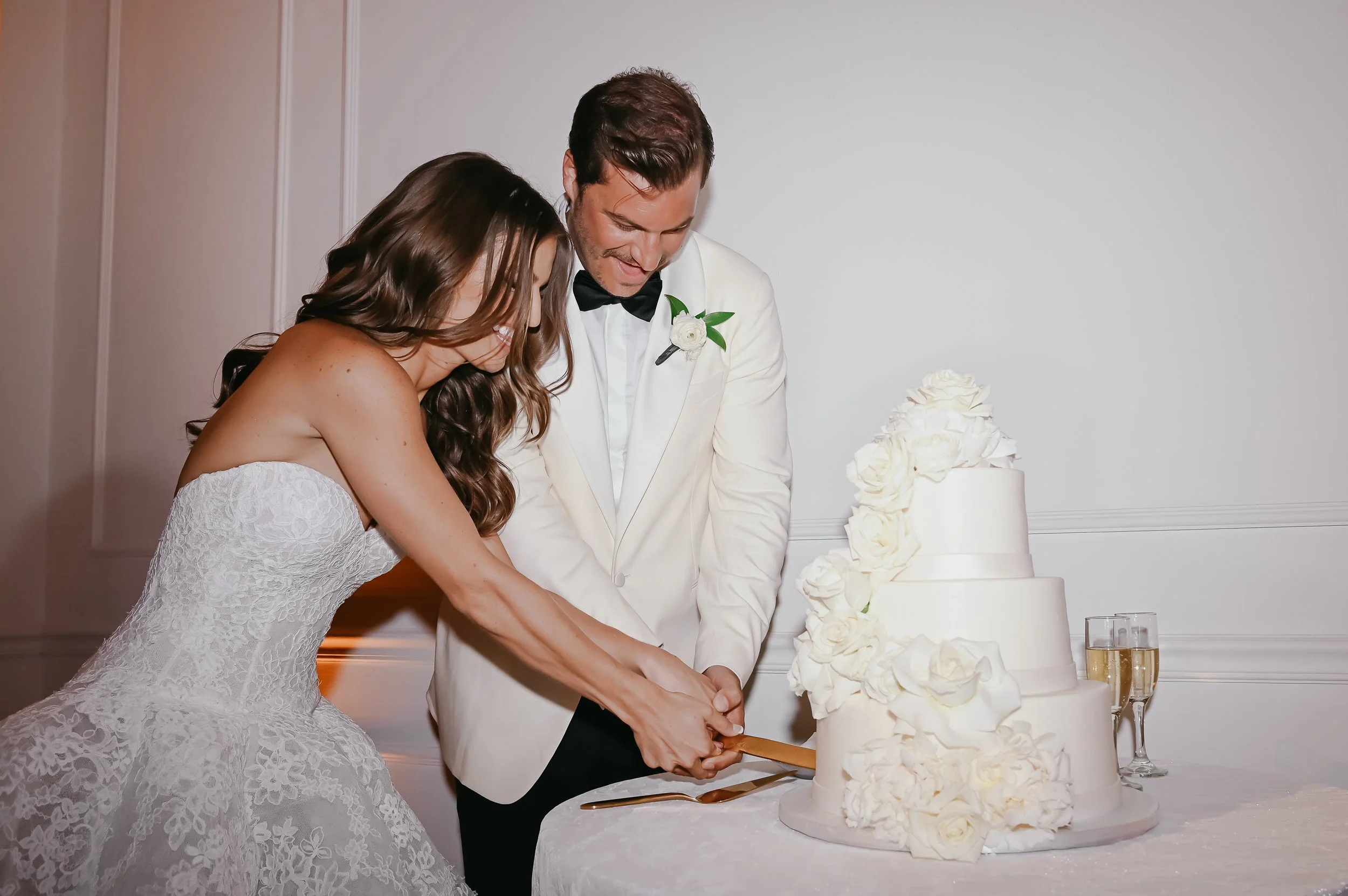 A bride and groom cutting a white wedding cake with floral decorations, standing in a room with white wall paneling.