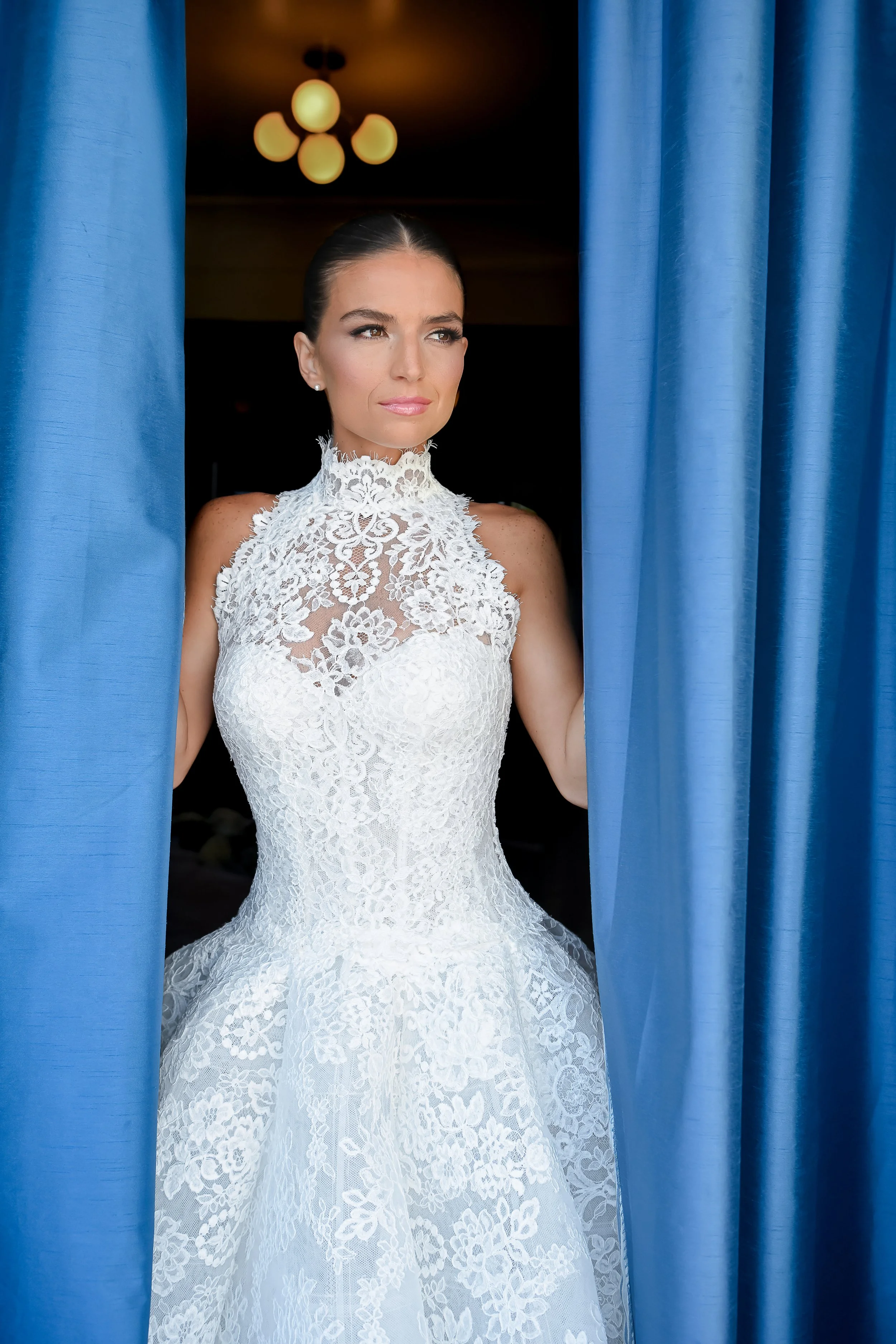 A woman in a white lace wedding dress standing between blue curtains, looking to the side with a serene expression.