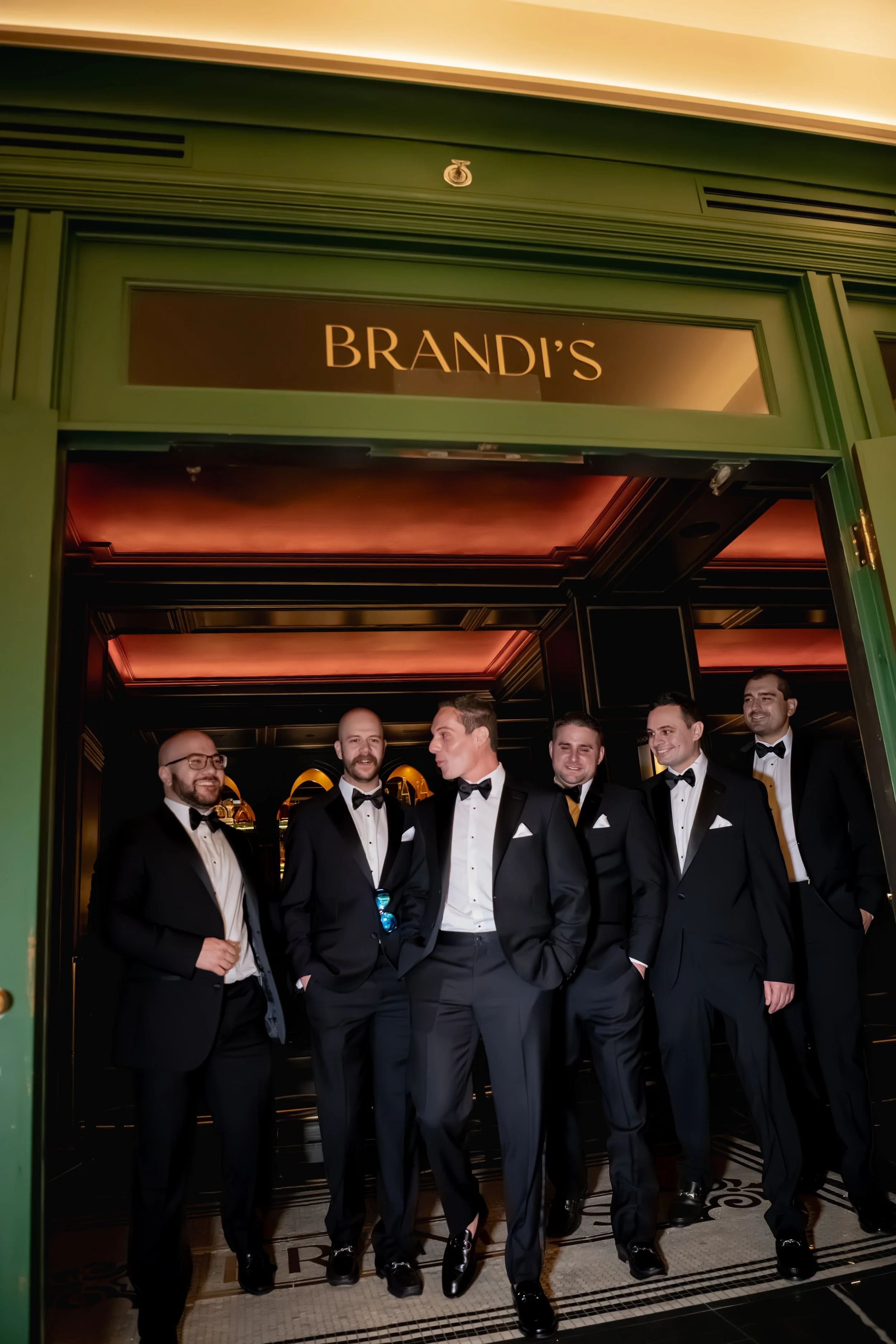Group of men in tuxedos standing inside a venue with a green frame and illuminated ceiling, with a sign above that reads 'BRANDI'S'.