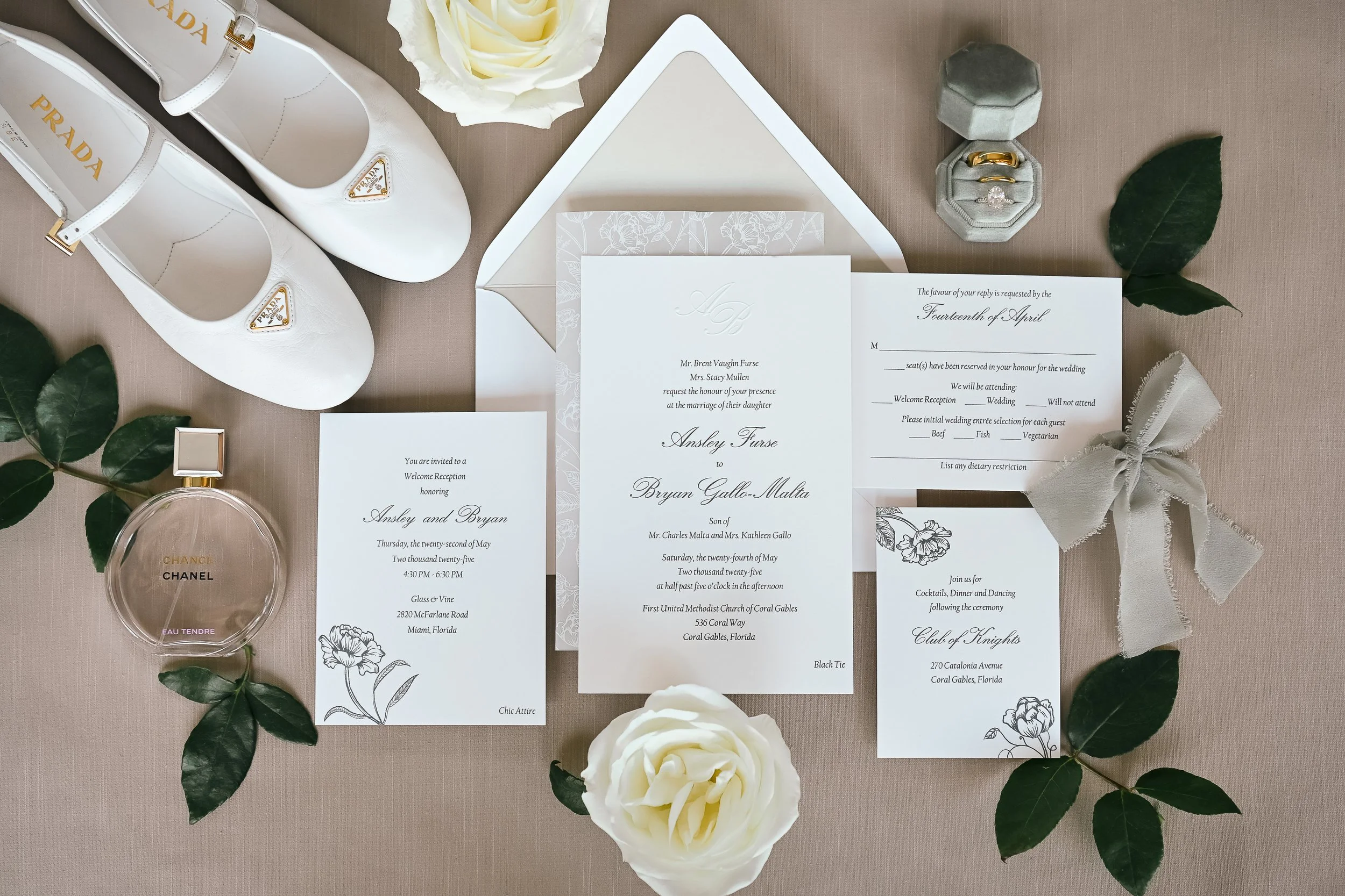 Wedding invitation suite with floral design, surrounded by white roses, green leaves, a Prada white shoe, perfume, and jewelry.