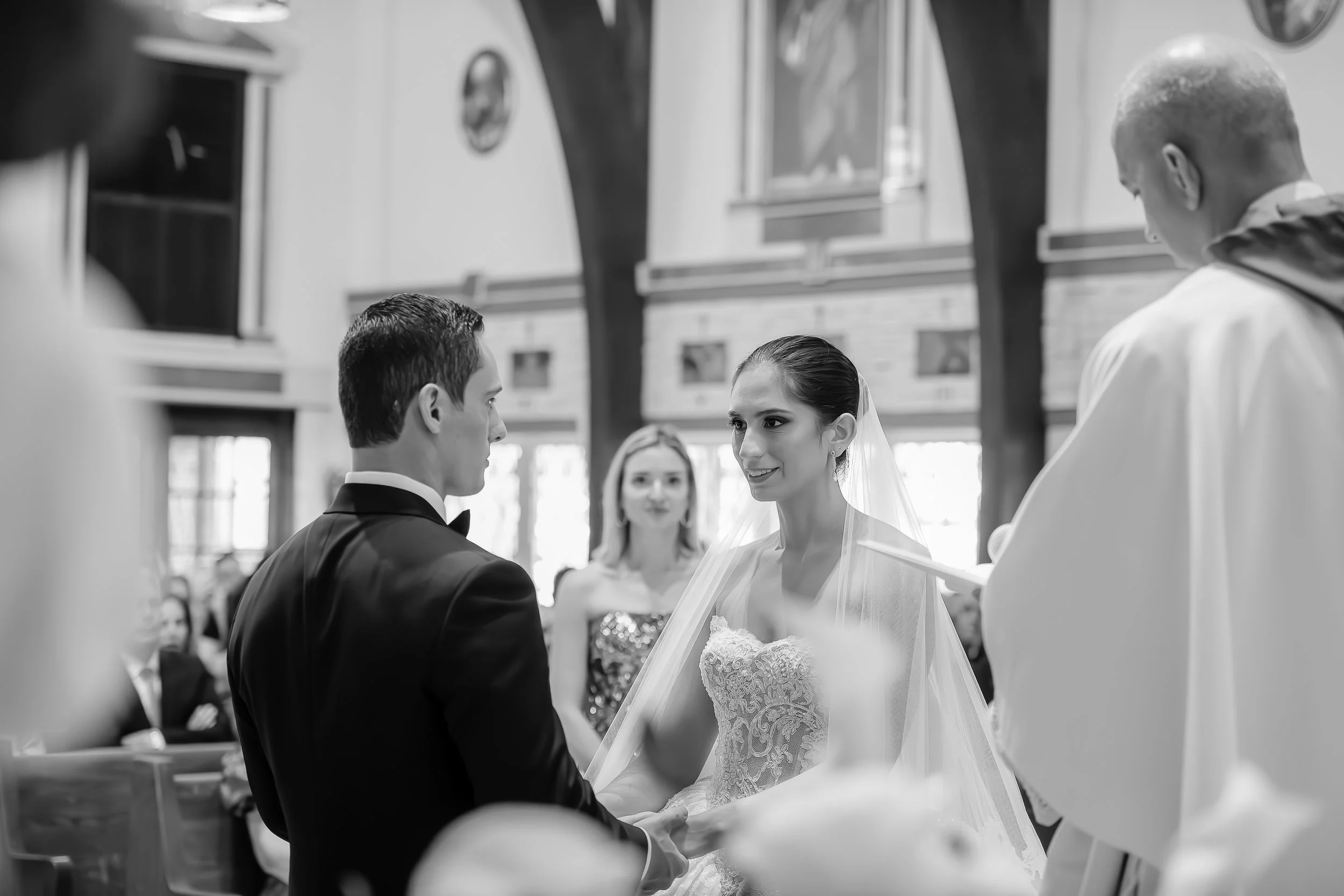 A black-and-white photo of a wedding ceremony inside a church. The bride and groom are facing each other, the bride in a lace wedding dress and veil, and the groom in a tuxedo. An officiant stands to the right, and a woman is visible in the backgroun
