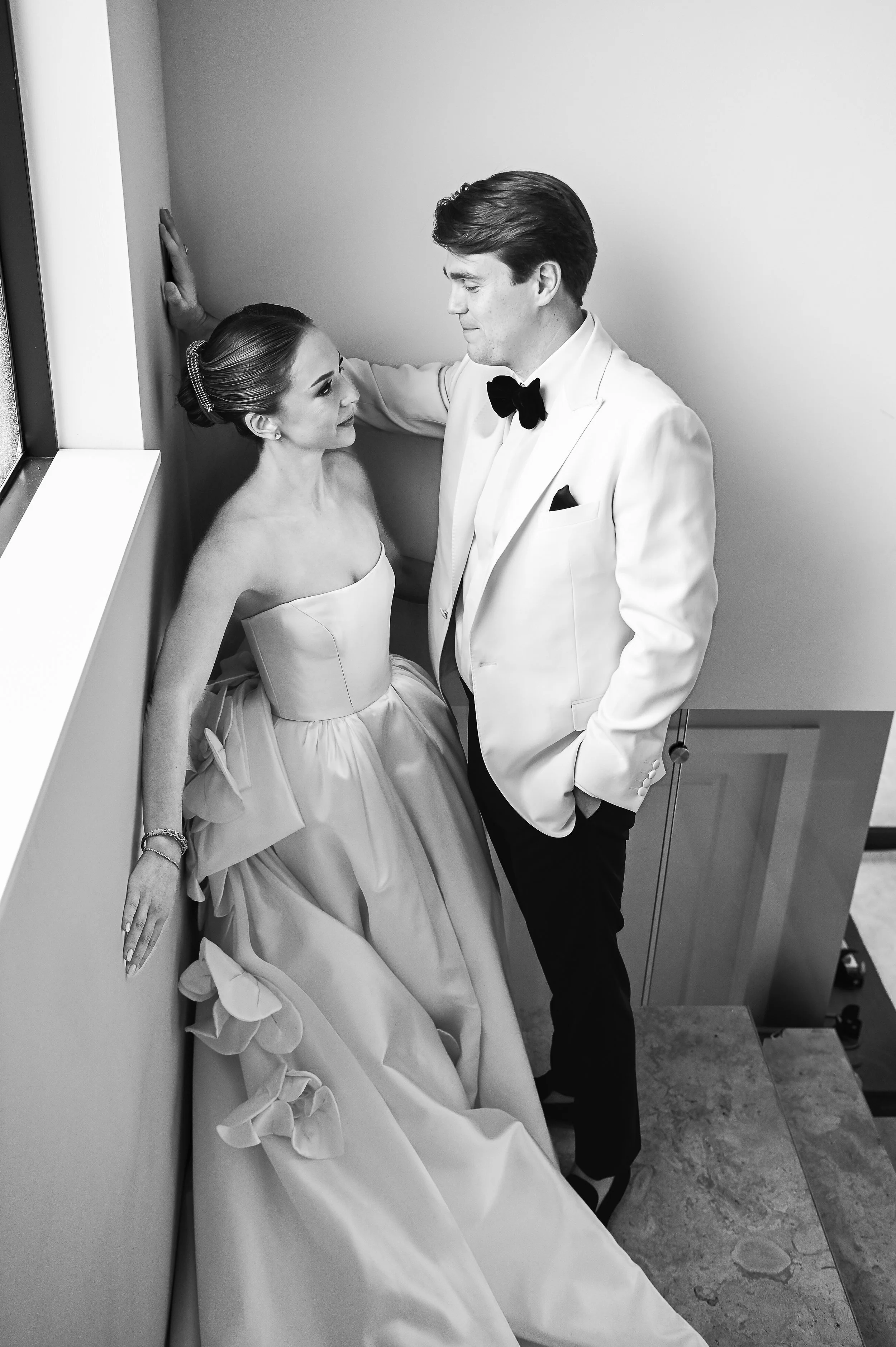 Black and white photo of a bride and groom in formal wedding attire standing in a corner, with the bride leaning against a wall and the groom looking at her, both with expressions of affection.