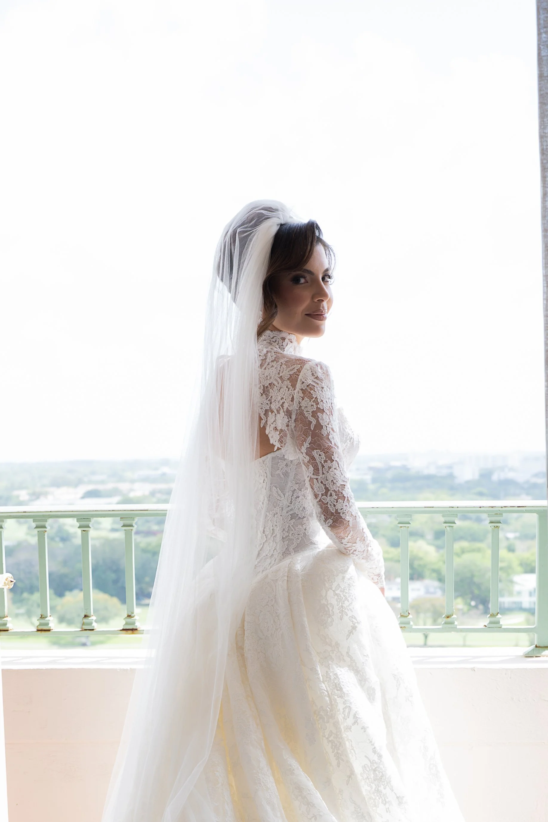 A bride with shoulder-length brown hair wearing a lace wedding dress and a veil, standing on a balcony with a view of a city and greenery in the background, looking over her shoulder.