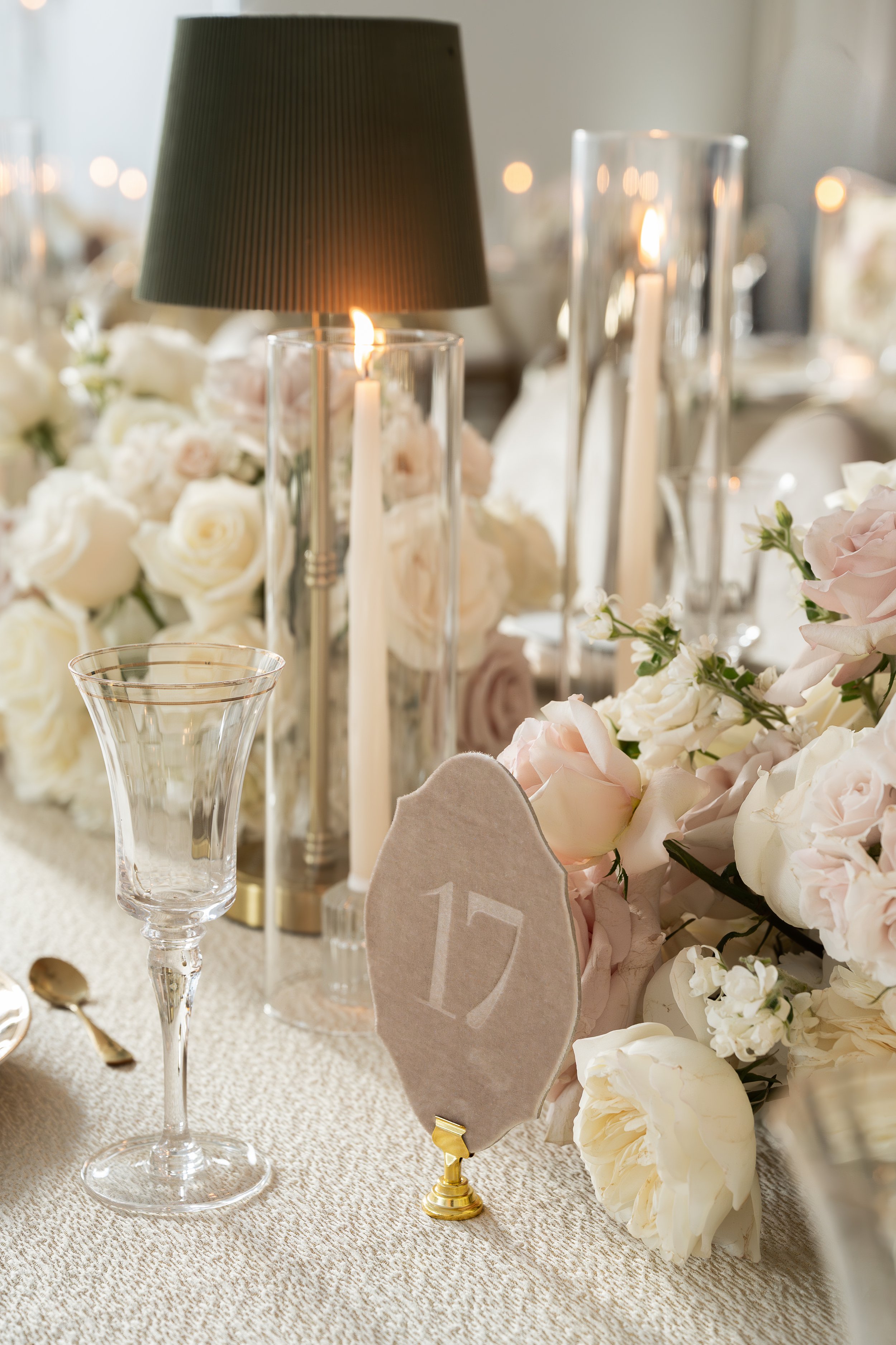 Elegant table setup with white and blush roses, tall candles, a grey lampshade with a candle inside, clear glassware, golden cutlery, and a beige tablecloth for a wedding or formal event.