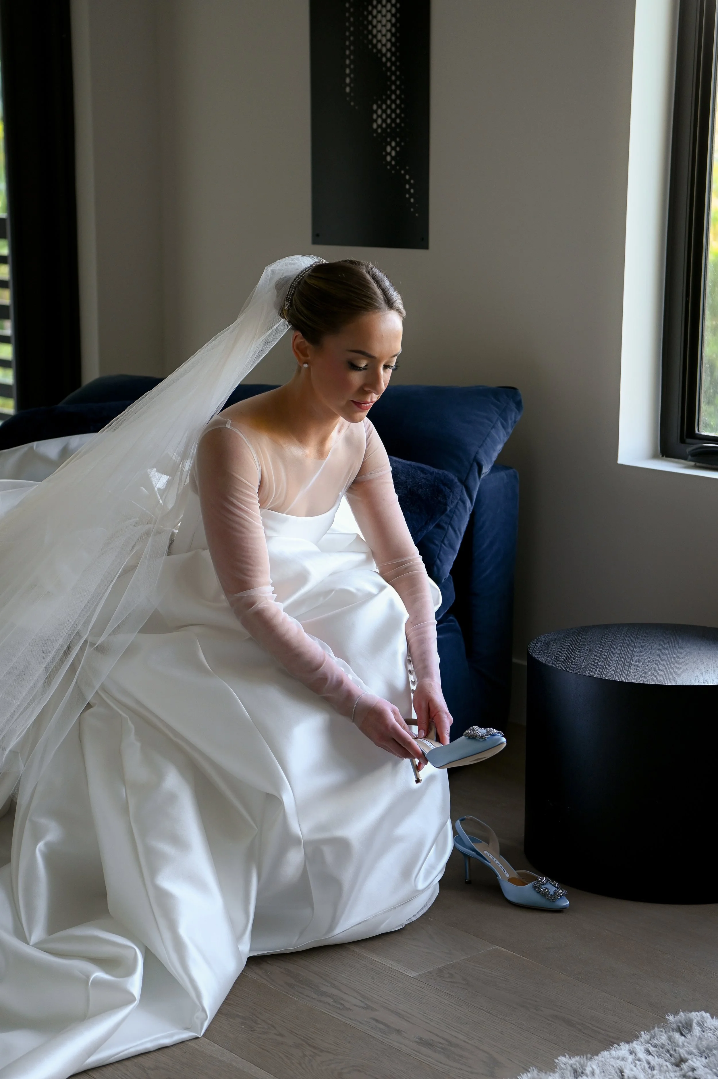 Bride in a wedding dress sitting on the floor, holding a shoe and looking down, with a veil draped over her shoulders, in a room with large windows and a dark table beside her.