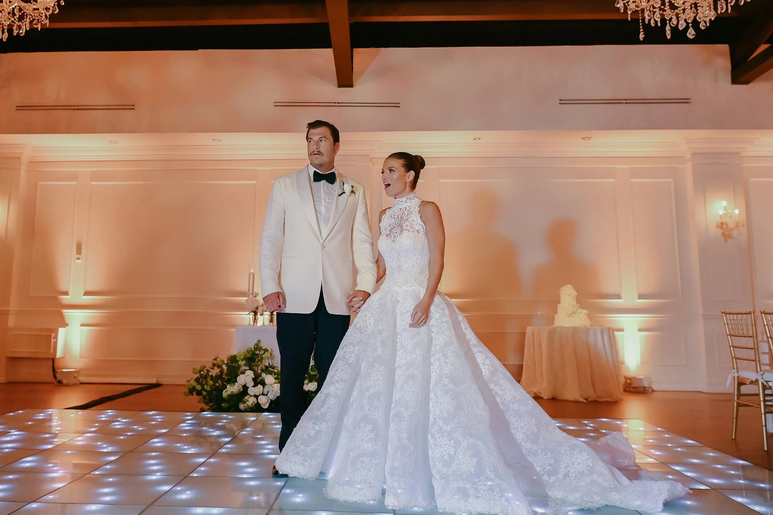 A bride and groom standing together during their wedding reception, holding hands, in an elegant ballroom with chandeliers, pastel-colored lighting, and a decorated table with a wedding cake in the background.