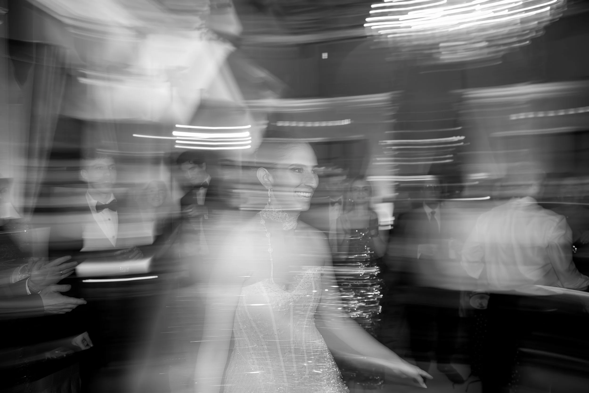 Black and white photo of a woman in a wedding dress smiling, with blurred guests and swirling lights in the background during a celebration or wedding reception.
