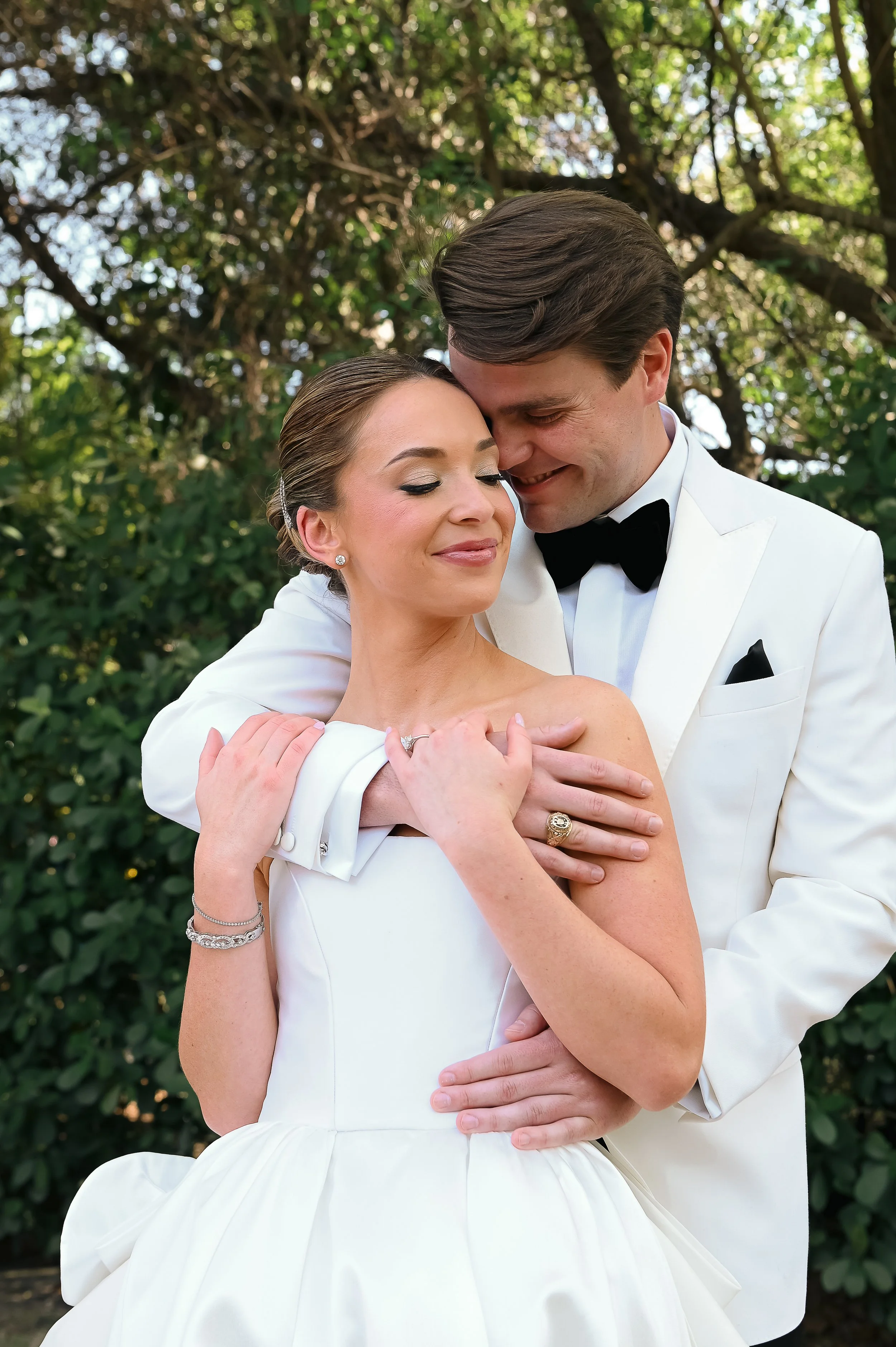 A bride and groom embracing outdoors, both smiling with eyes closed, surrounded by green trees.