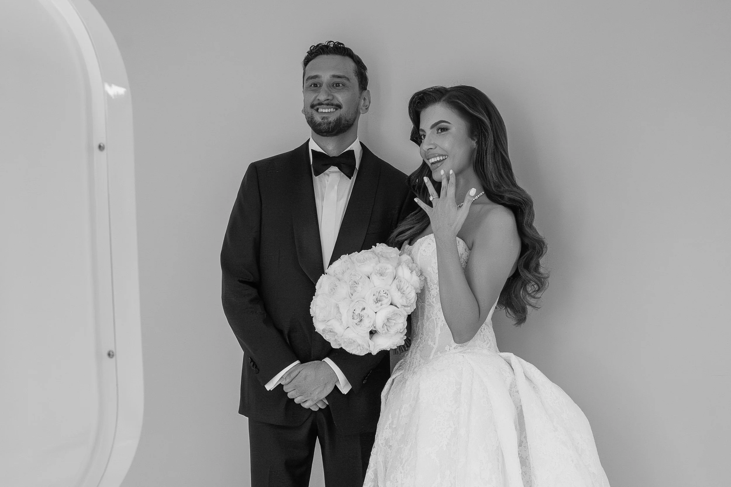 Black and white photo of a smiling bride and groom taking a selfie together at their wedding, with the bride holding a bouquet of white roses.