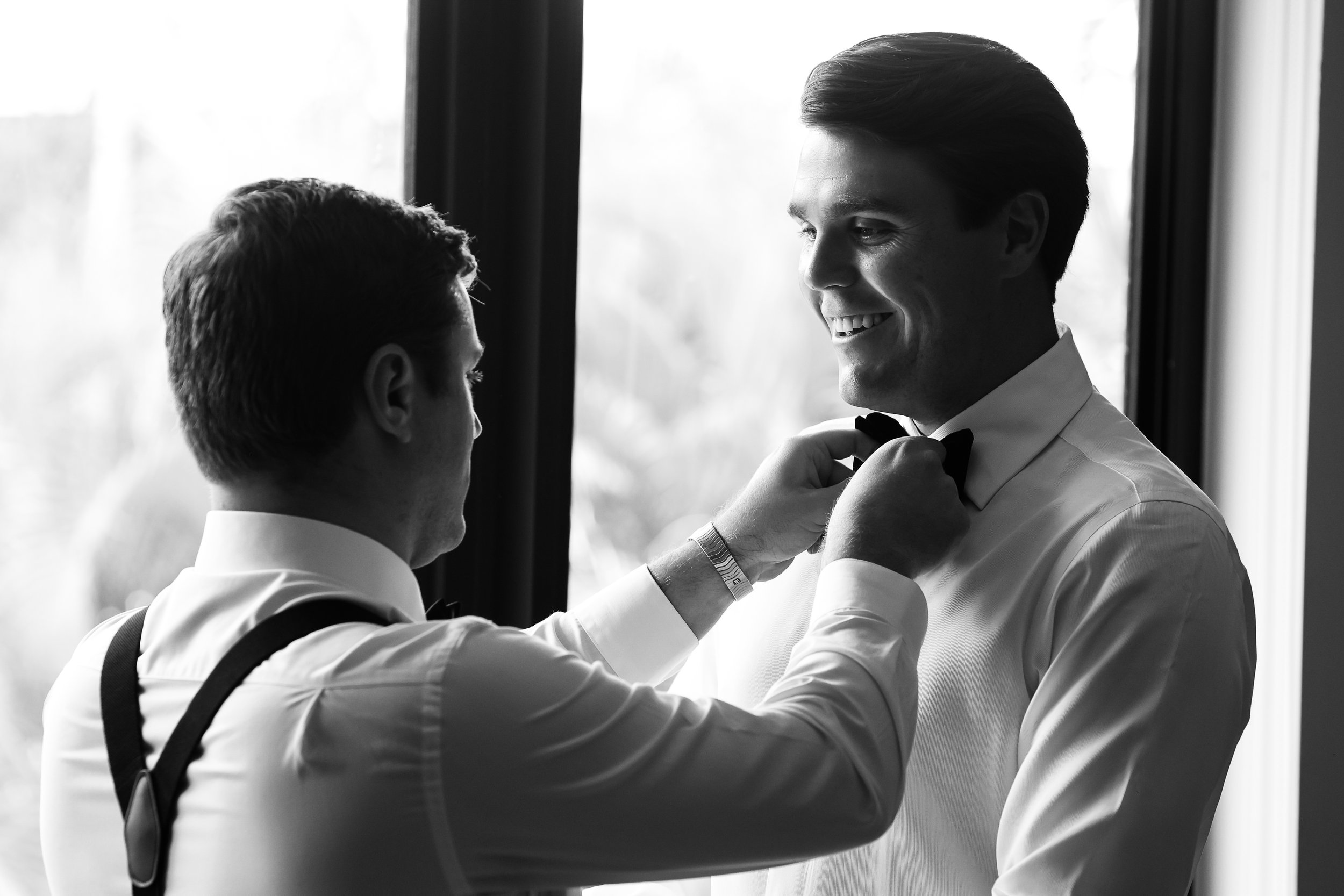 Two men with light skin in formal attire, one helping the other adjust a bow tie near a window.