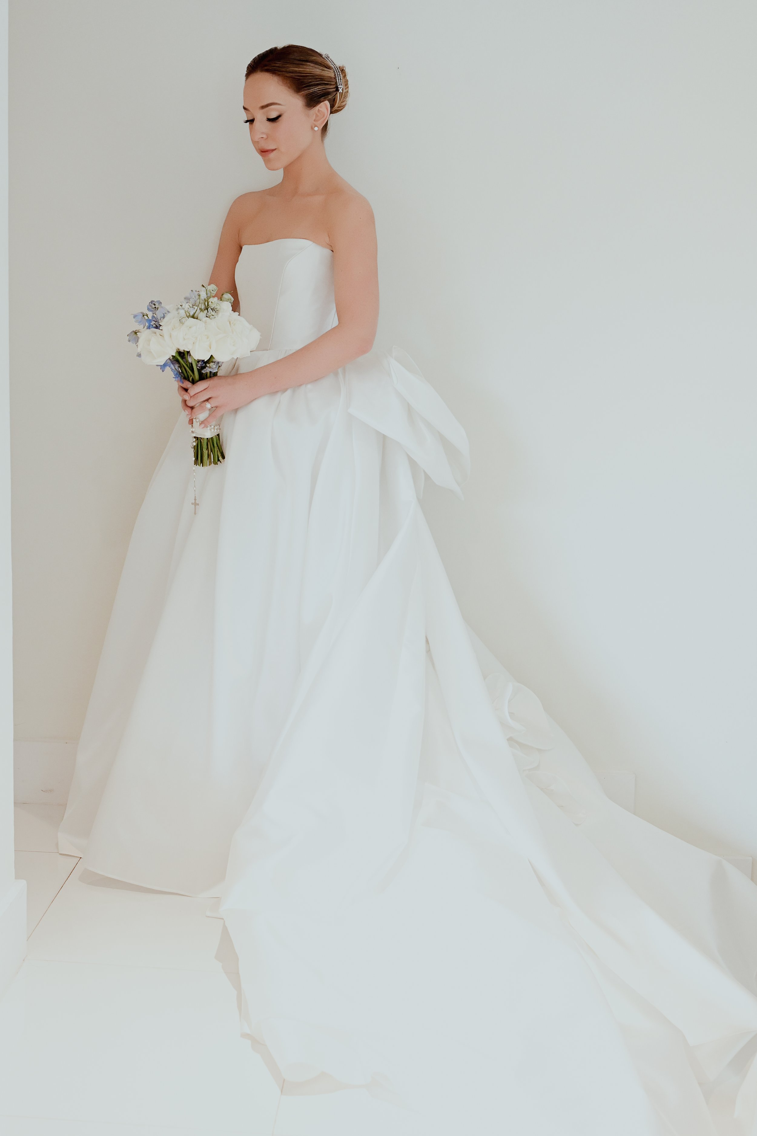A young woman in a white wedding dress holding a bouquet of flowers. She has her hair styled in an elegant updo and is standing against a plain white background.