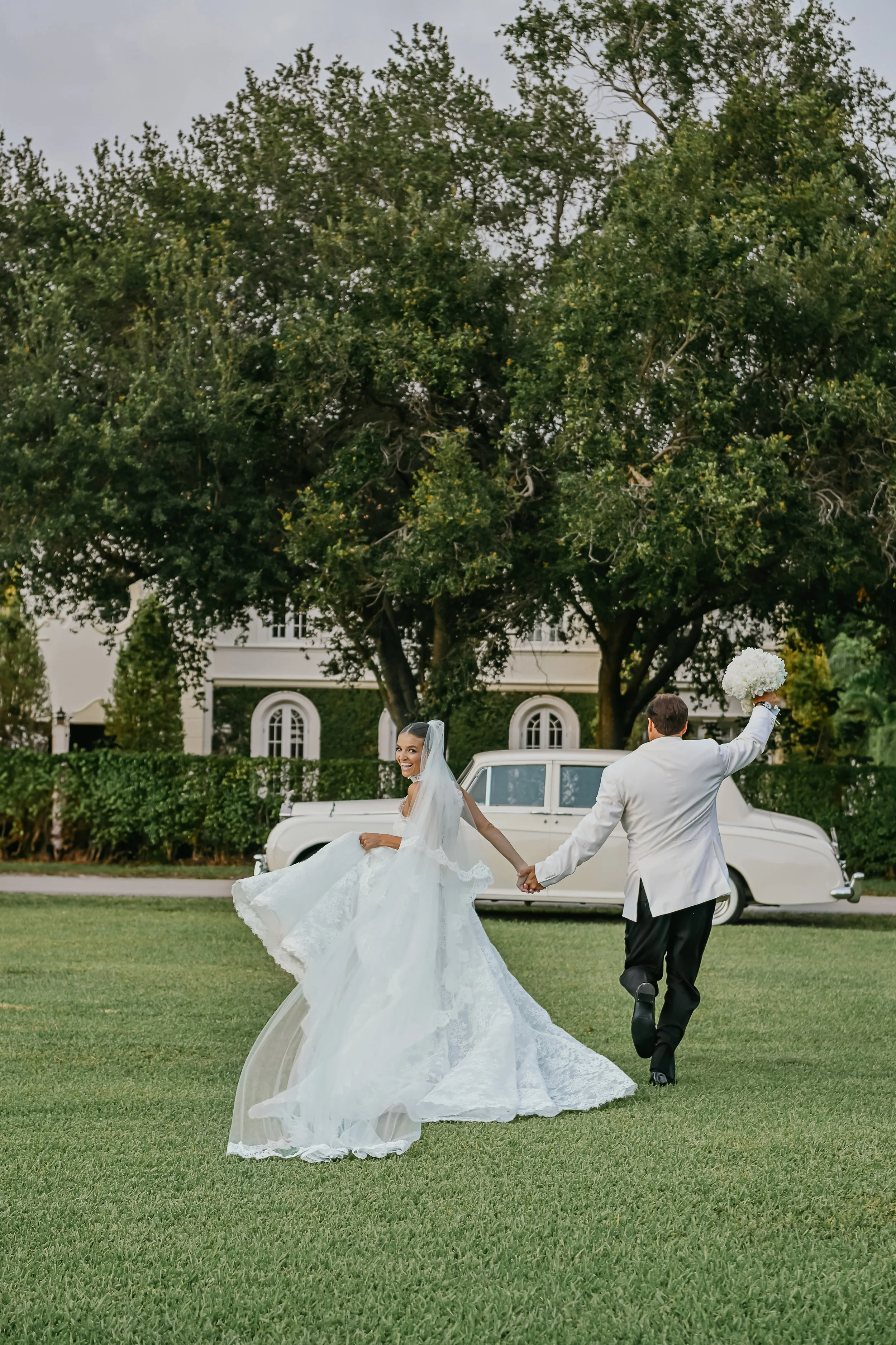 Bride and groom holding hands, walking on a lawn outdoors, with large trees and a white building in the background. The bride is smiling and wearing a white wedding dress, while the groom is dressed in a white jacket and black pants, holding a bouquet of white flowers.