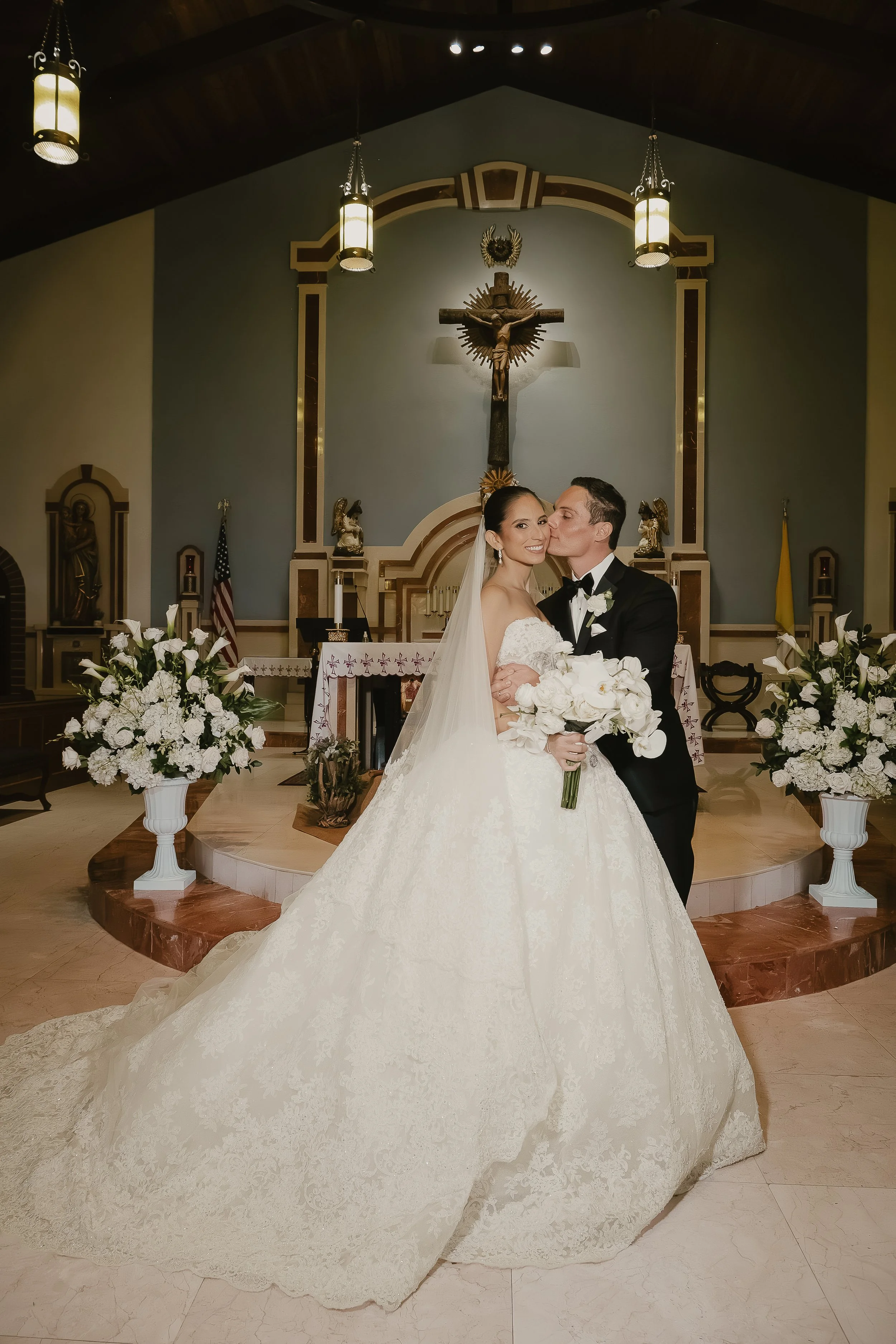 A bride and groom standing together inside a church, with the bride holding a bouquet of white flowers. The church has an altar behind them, with religious statues, a large crucifix, and floral arrangements on either side. The bride is wearing a whit