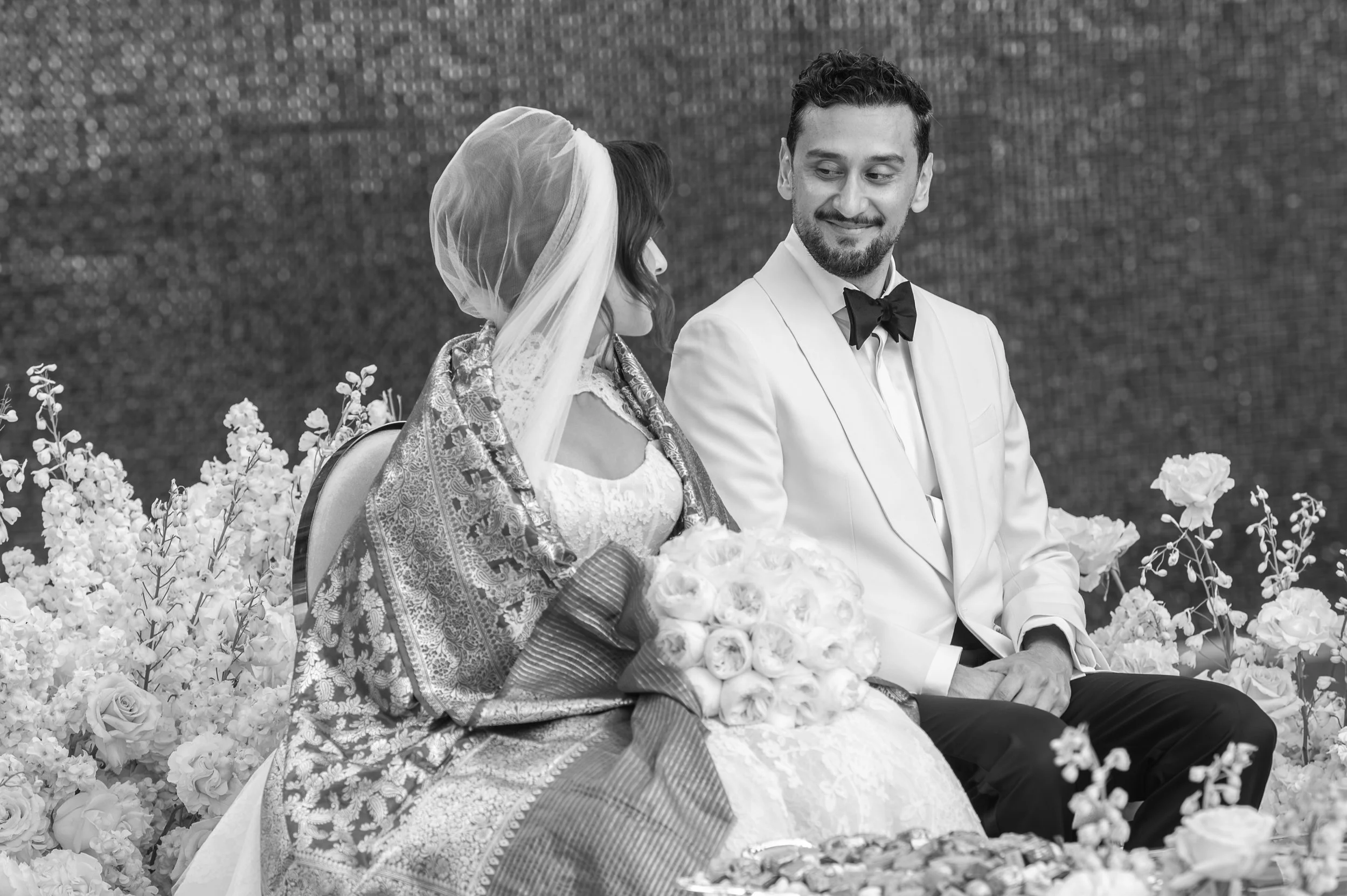 A black and white photo of a wedding scene with a bride and groom sitting together among flowers, the bride wearing a lace dress and a head covering, holding a bouquet, and the groom in a white suit with a bow tie, both looking at each other with smi
