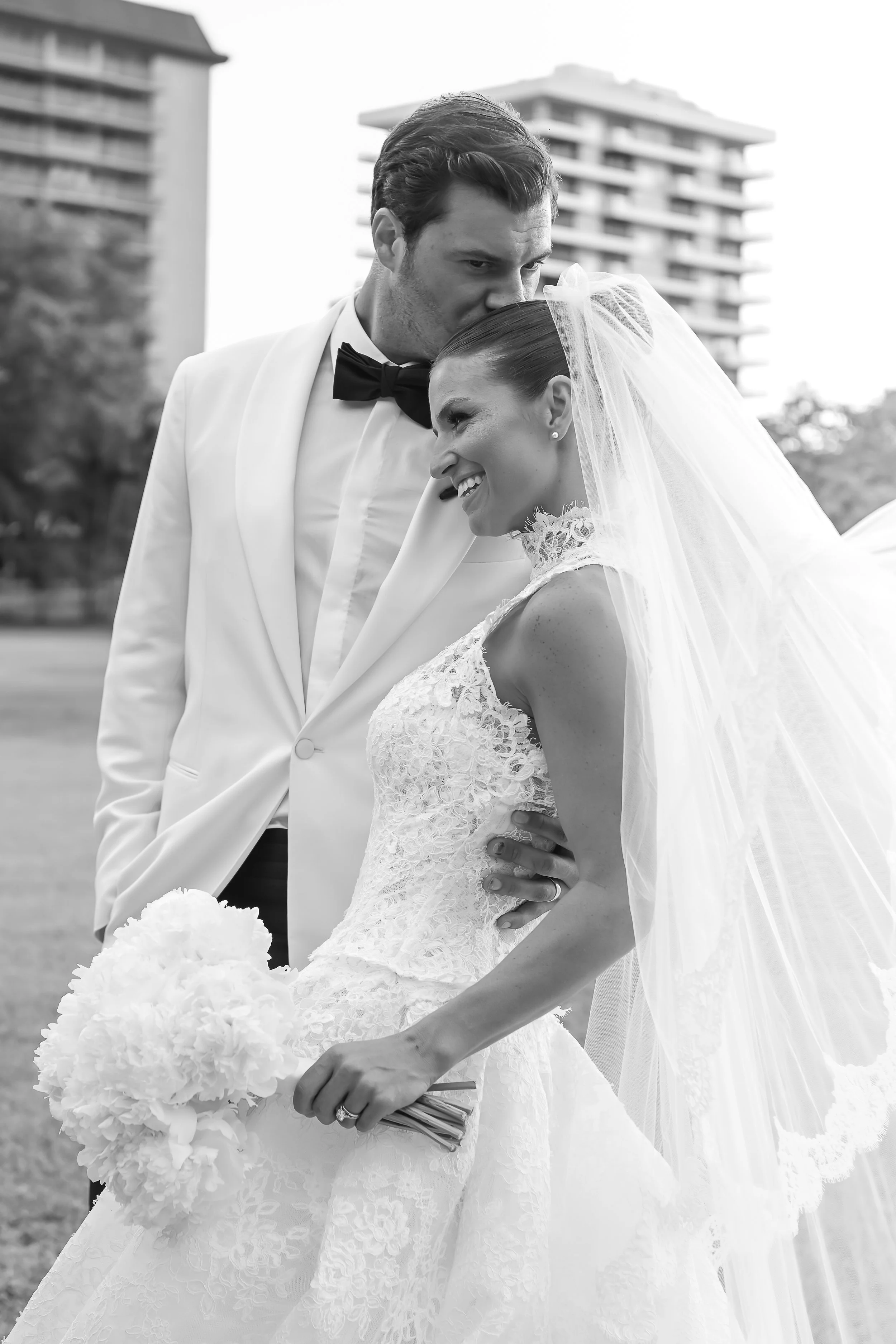 Black and white photograph of a bride and groom on their wedding day outdoors. The groom is in a white tuxedo with a black bow tie, leaning to kiss the bride on her forehead. The bride is smiling, wearing a lace wedding gown and veil, holding a bouqu