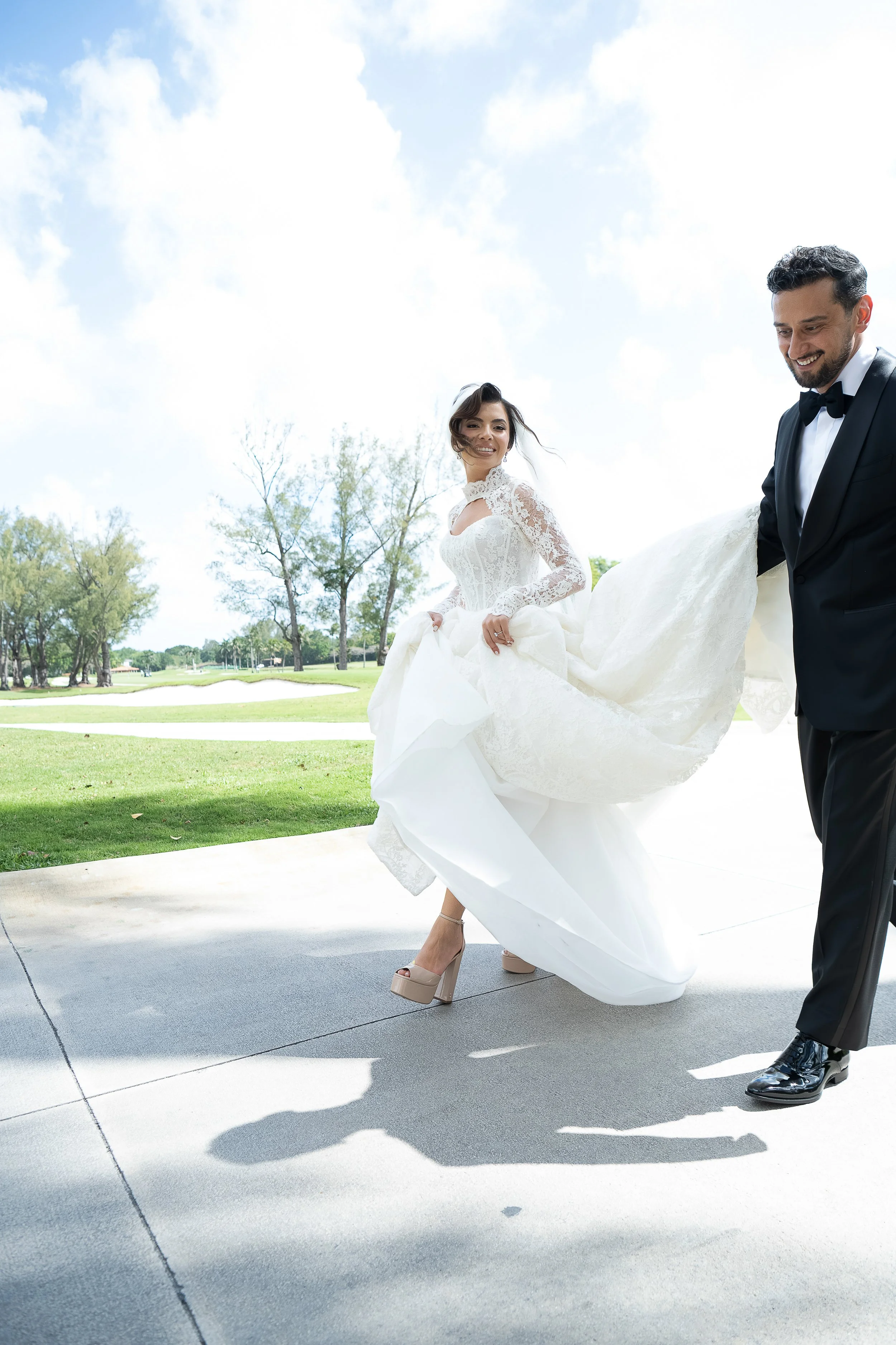 Bride in a white wedding dress with lace sleeves and high heels, laughing and holding her dress as she walks with a groom in a tuxedo with a bow tie, outdoors with trees and a blue sky.