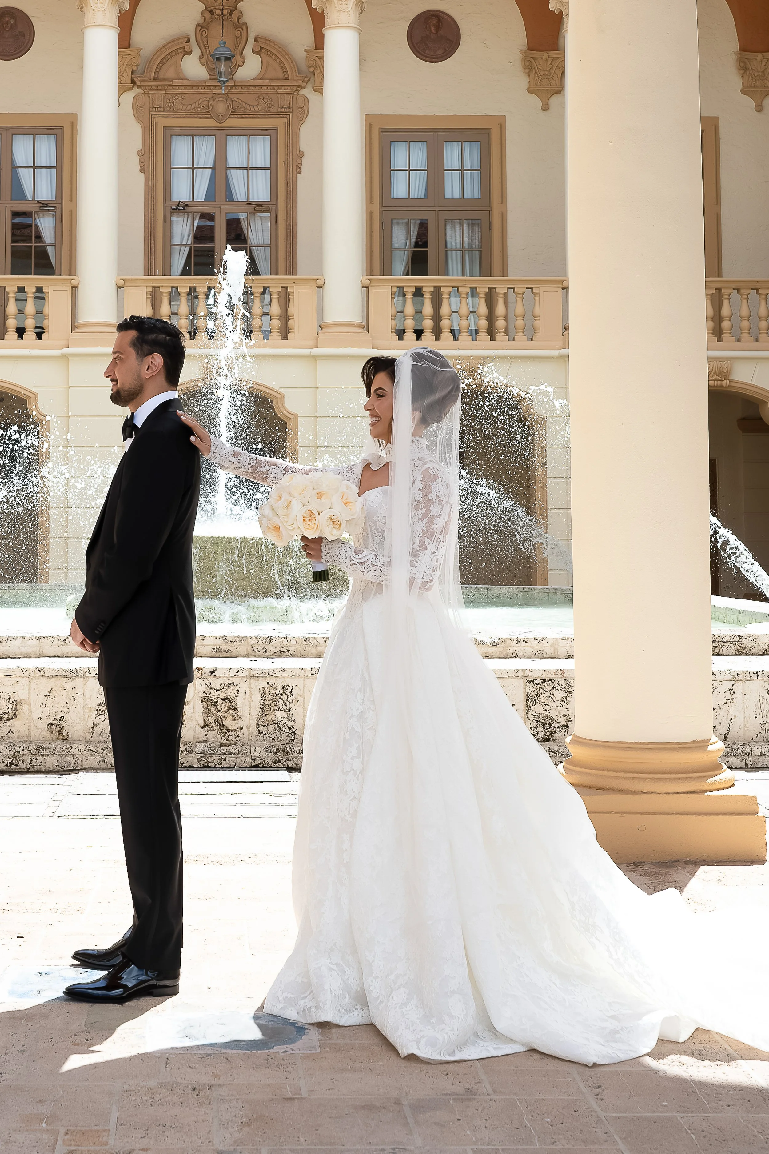 A bride in a white lace wedding gown and veil holding a bouquet of roses stands behind a groom in a black tuxedo. They are outdoors in front of a building with columns and ornate architectural details near a fountain.