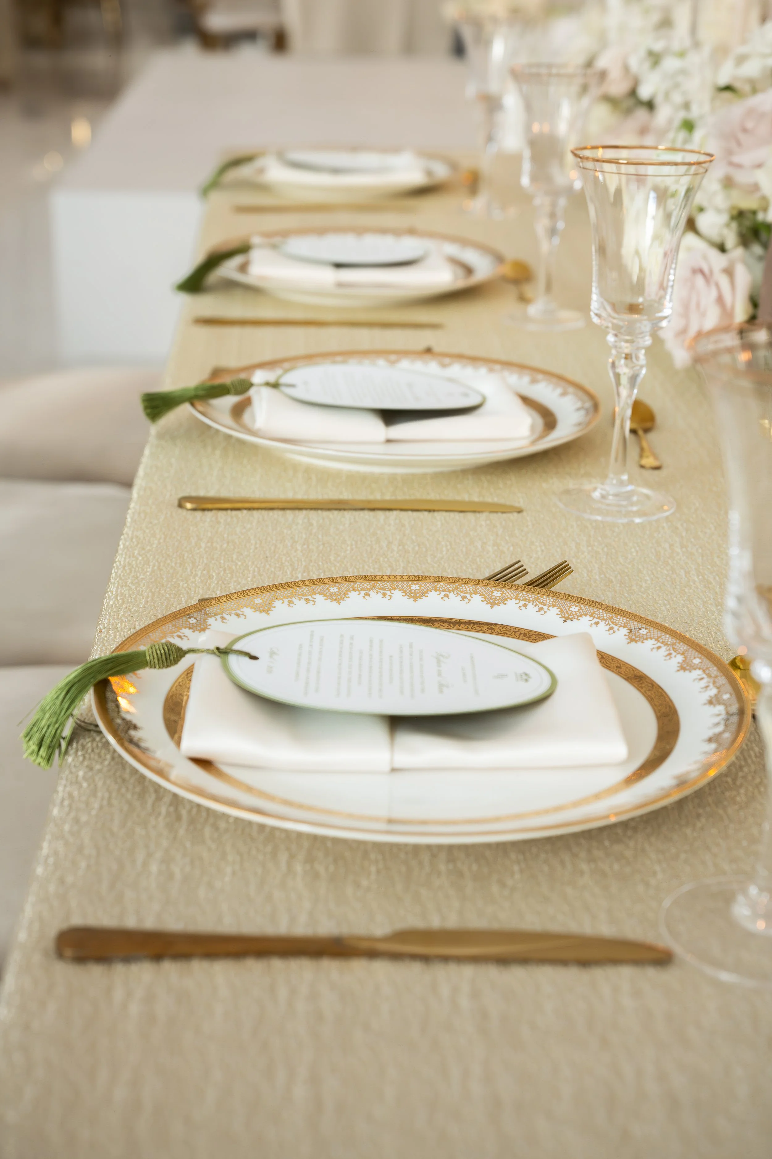A long banquet table with elegant place settings, including gold-rimmed china plates, white napkins, and individual menus on each plate. Green tassel napkin rings and gold utensils are arranged, with tall, clear glass wine goblets with gold rims plac
