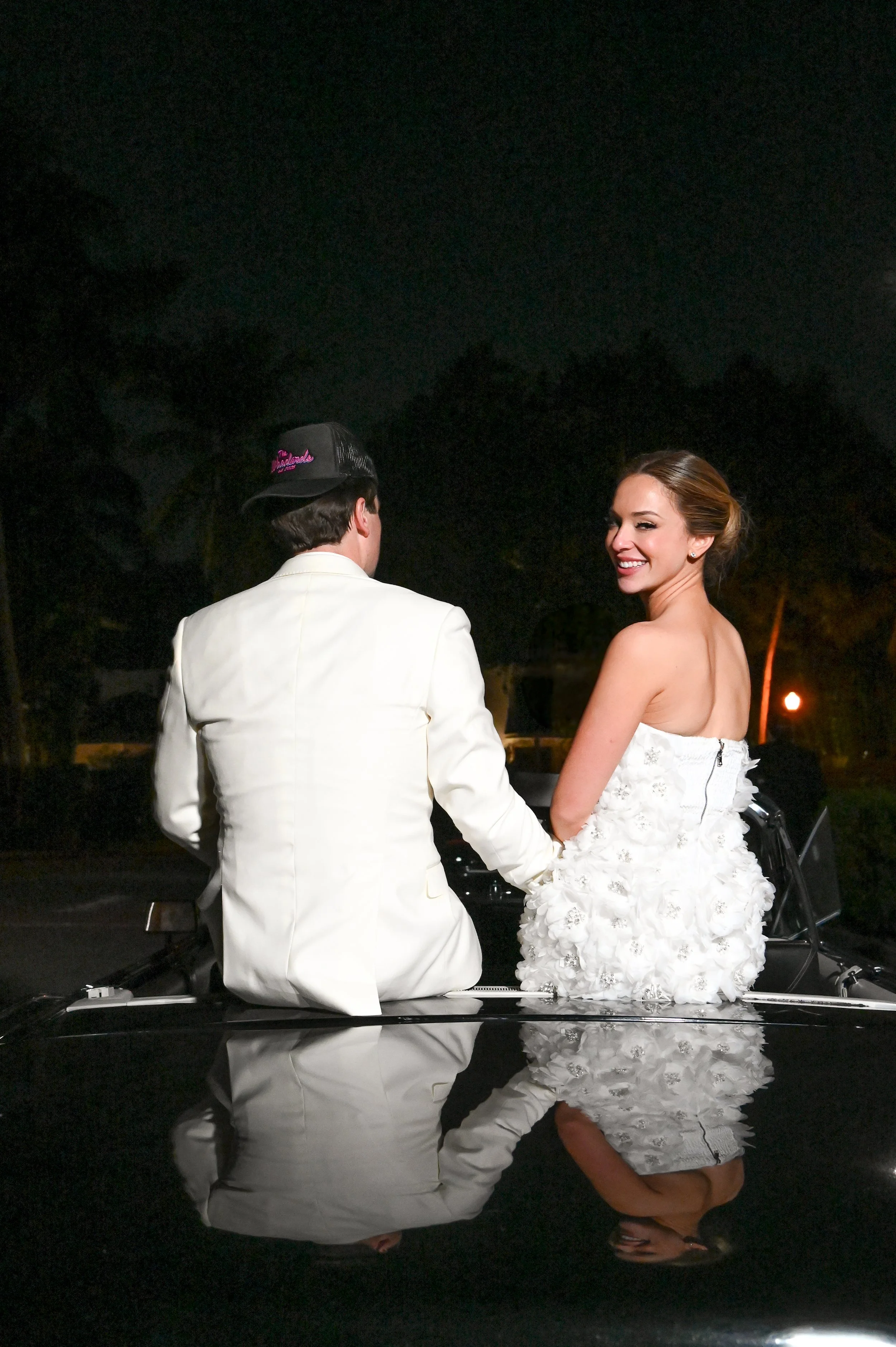 A couple dressed in wedding attire smiling and holding hands outdoors at night, with the woman's wedding dress visible and a reflection of the couple on a shiny surface.