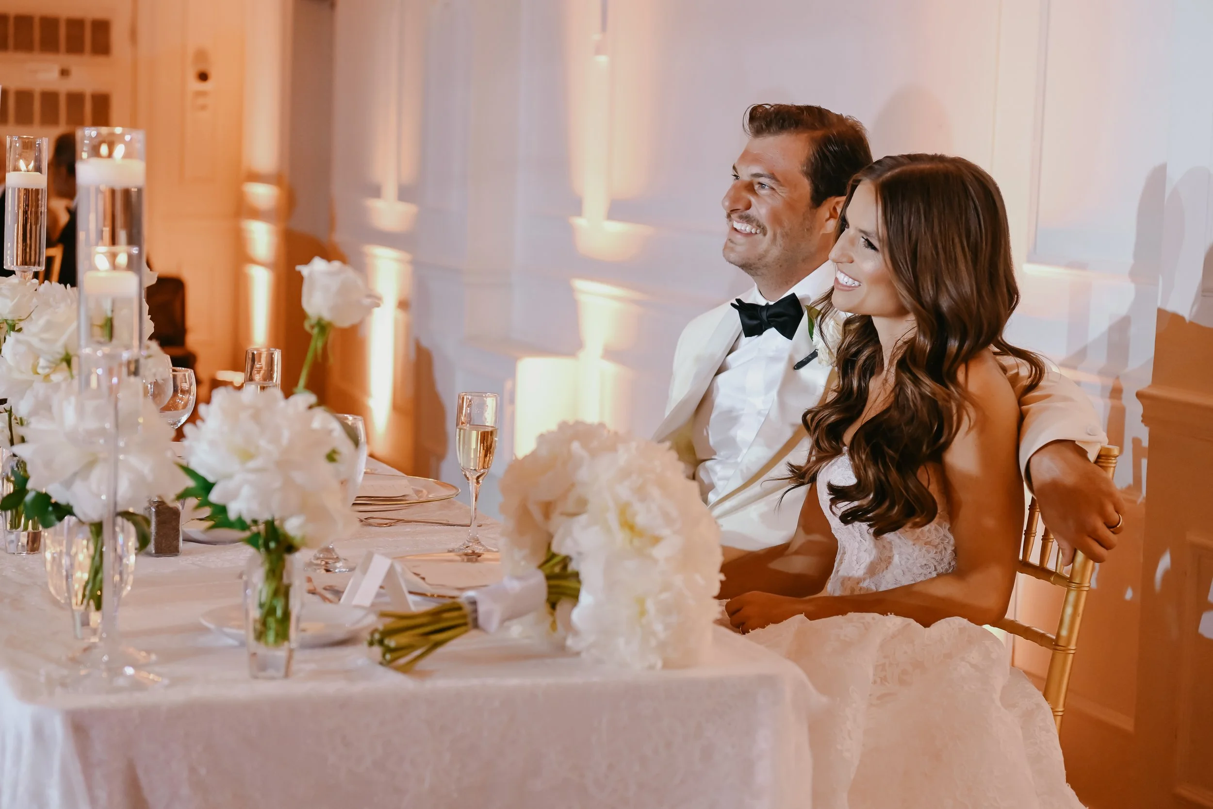 A bride and groom smiling and sitting at their wedding reception table decorated with white flowers, candles, and champagne glasses. The groom is wearing a tuxedo with a black bow tie, and the bride has long wavy brown hair and is wearing a white lac