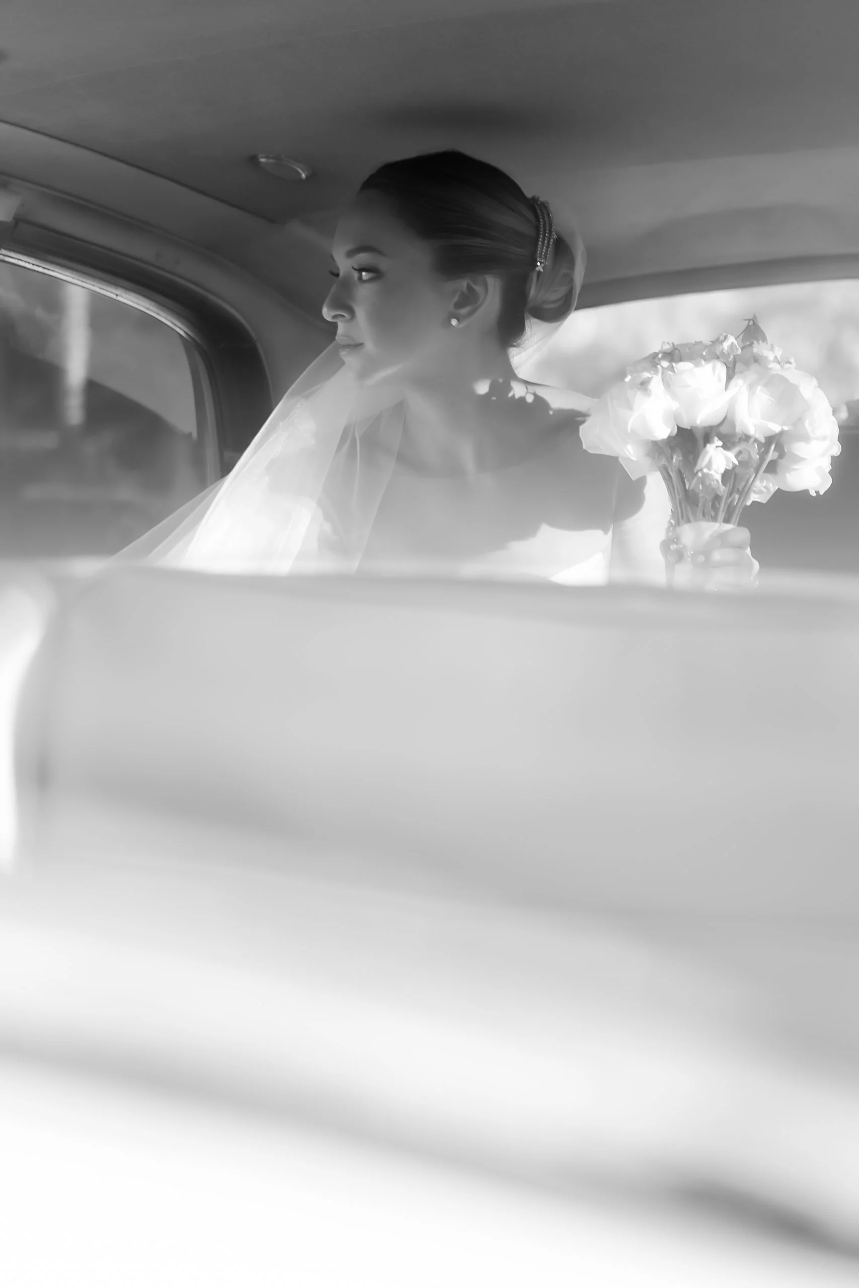 A bride inside a vehicle holding a bouquet of flowers, looking out the window in a black and white image.