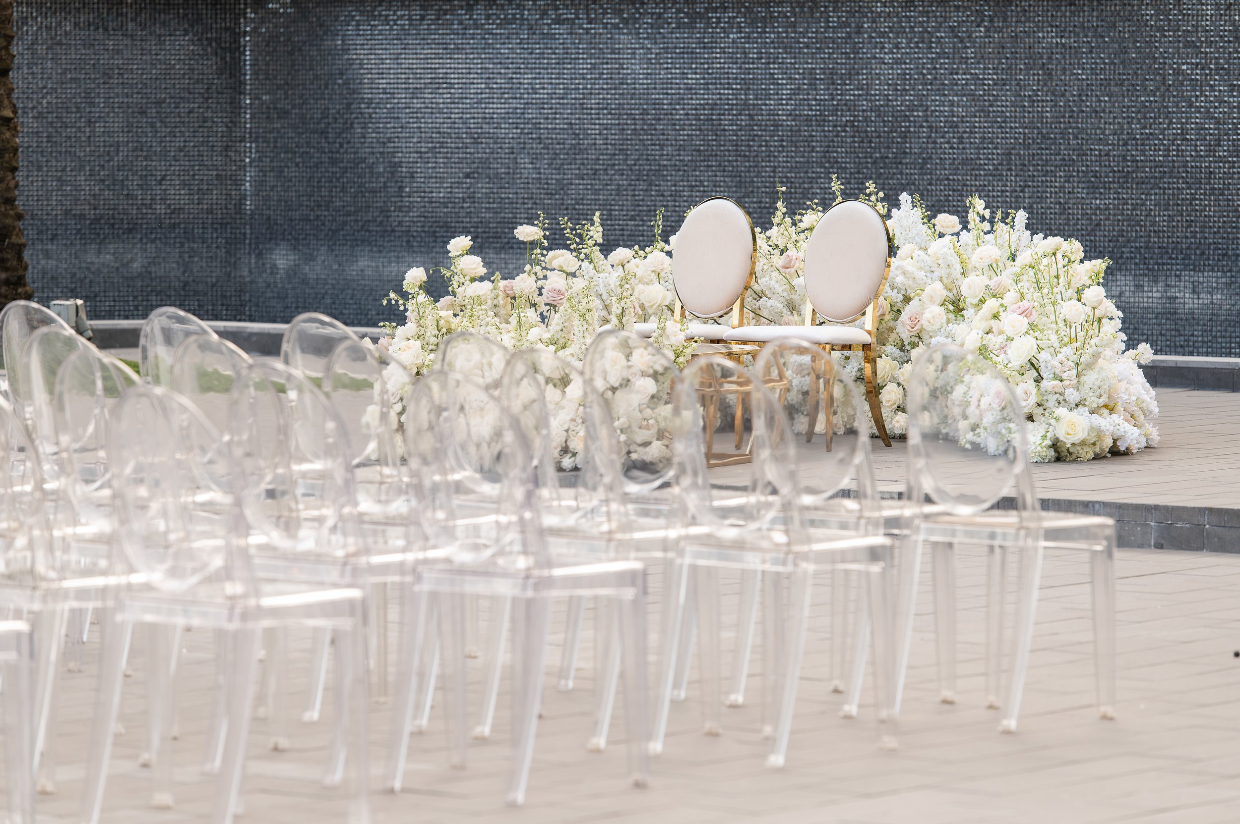 Elegant outdoor wedding setup with two white chairs and floral arrangements of white and pale pink roses and greenery on a stage, with transparent ghost chairs arranged in rows in front of the stage.