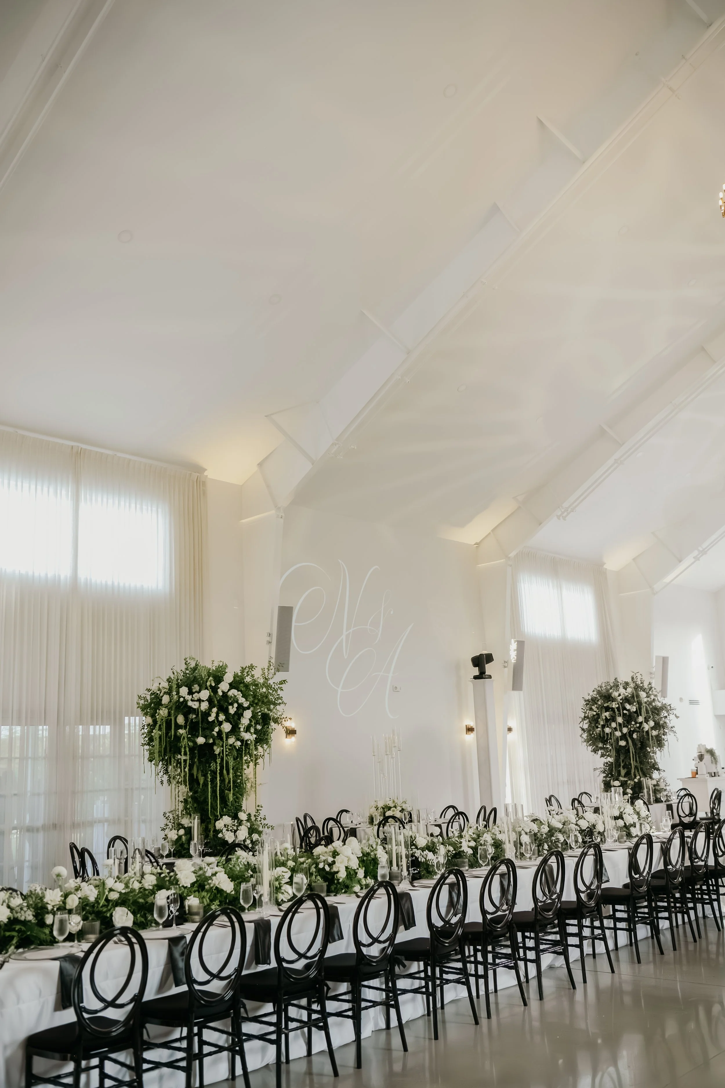Elegant banquet hall decorated with white flowers and greenery, set for a formal event with black chairs around long tables, tall windows with white curtains, and soft lighting.