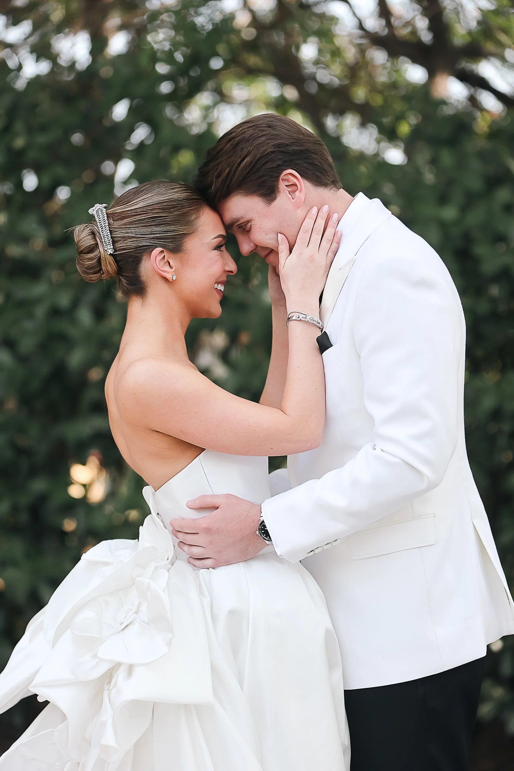 A bride and groom are facing each other closely, touching foreheads and smiling. The bride is wearing a strapless white wedding dress with a large bow at the back and has her hair styled in an elegant updo with a rhinestone hair accessory. The groom is dressed in a white tuxedo and has his hands gently placed on the bride's waist. They are outdoors with a blurred background of green foliage.