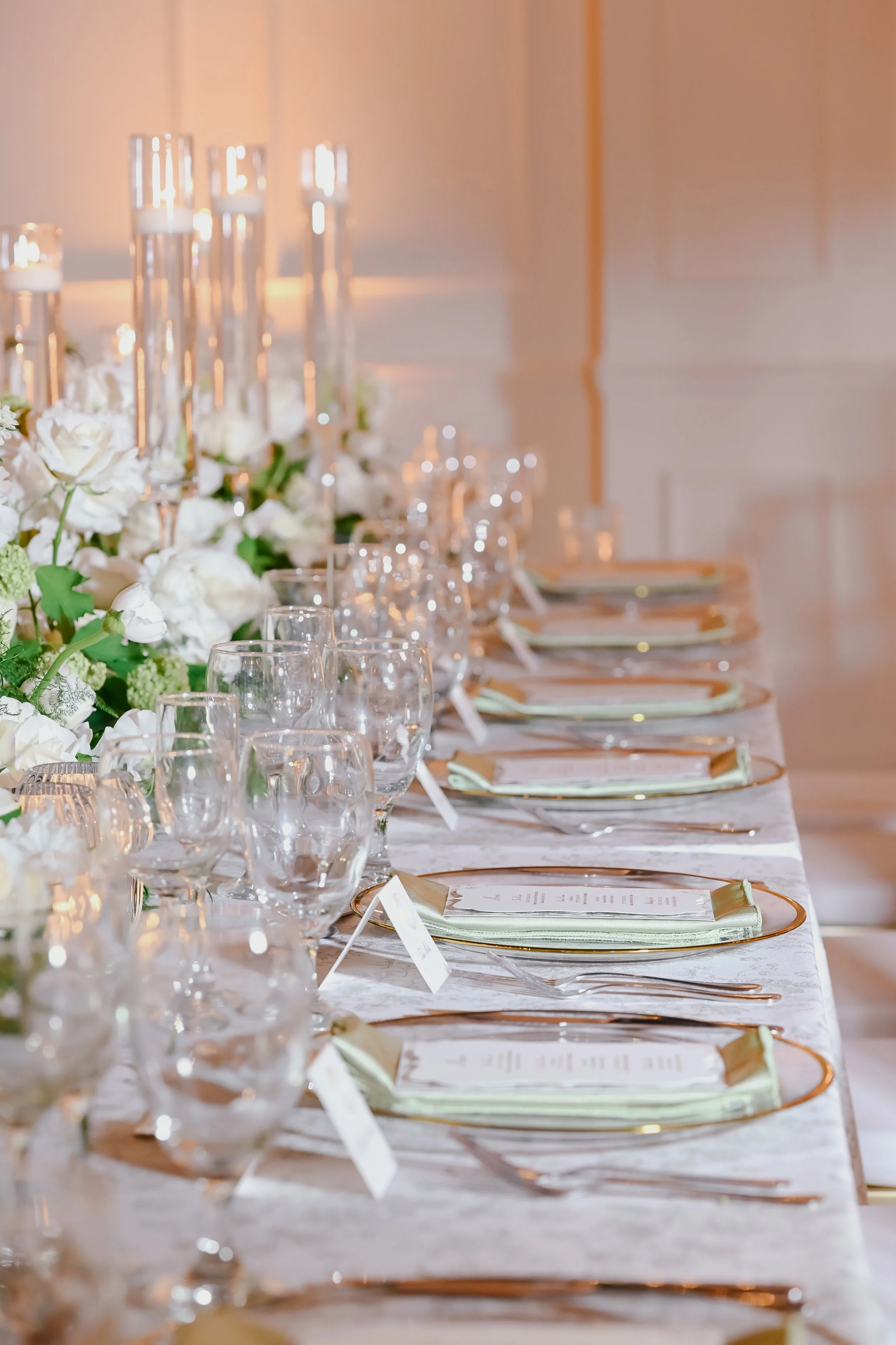 Elegant banquet table set with glassware, floral arrangements, gold-rimmed plates, silverware, and menu cards under candlelight.