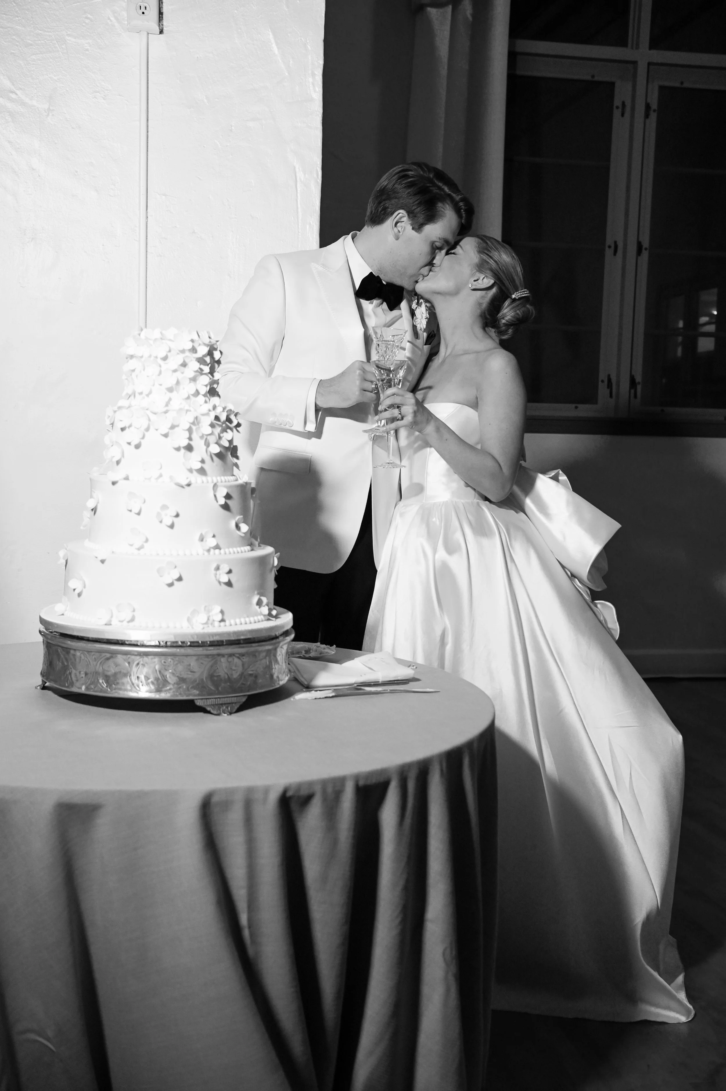 Black and white photo of a bride and groom sharing a kiss at their wedding reception, with a wedding cake nearby.